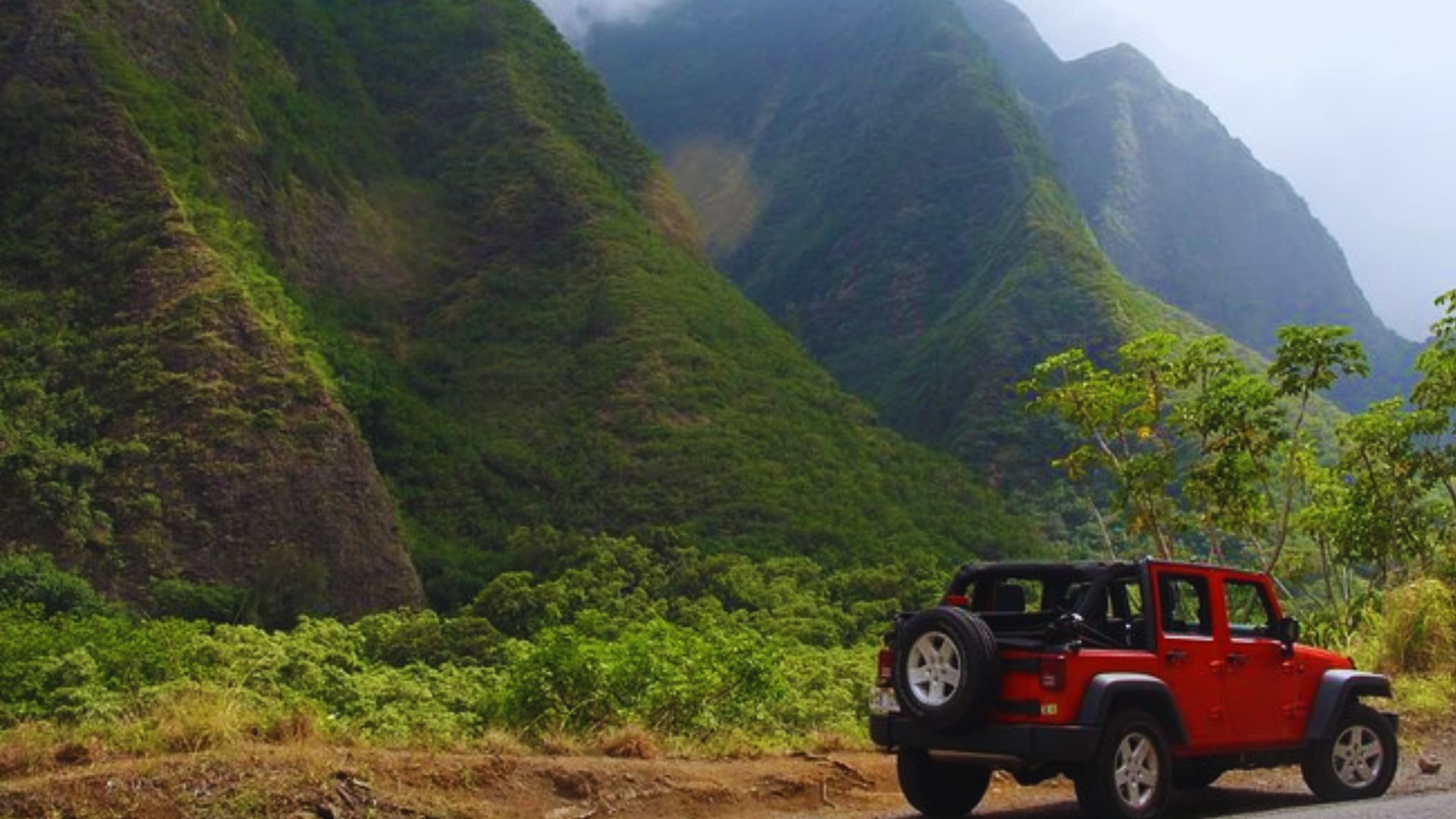 A red jeep navigates a winding mountain road surrounded by lush greenery and rocky terrain.