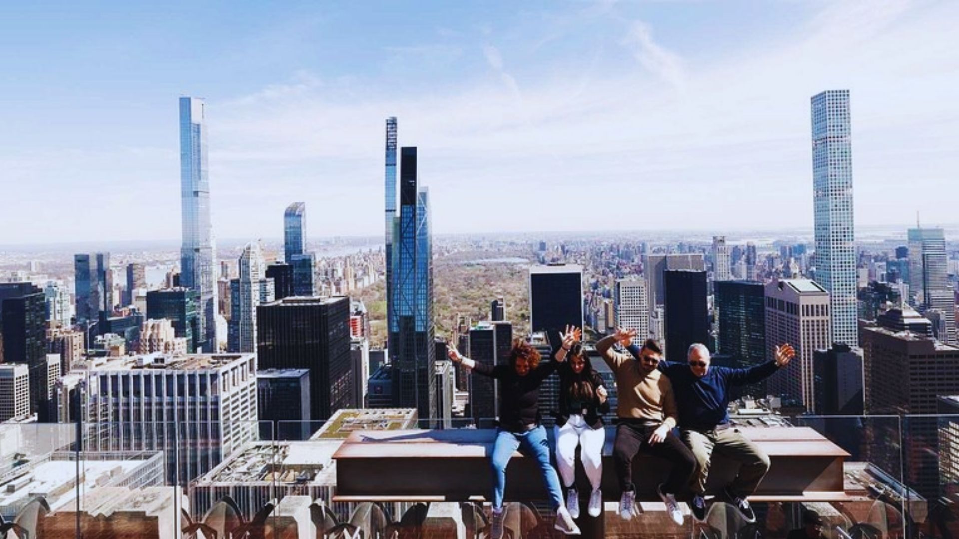 Three people sit on a ledge, gazing at the city skyline below them during a clear day.