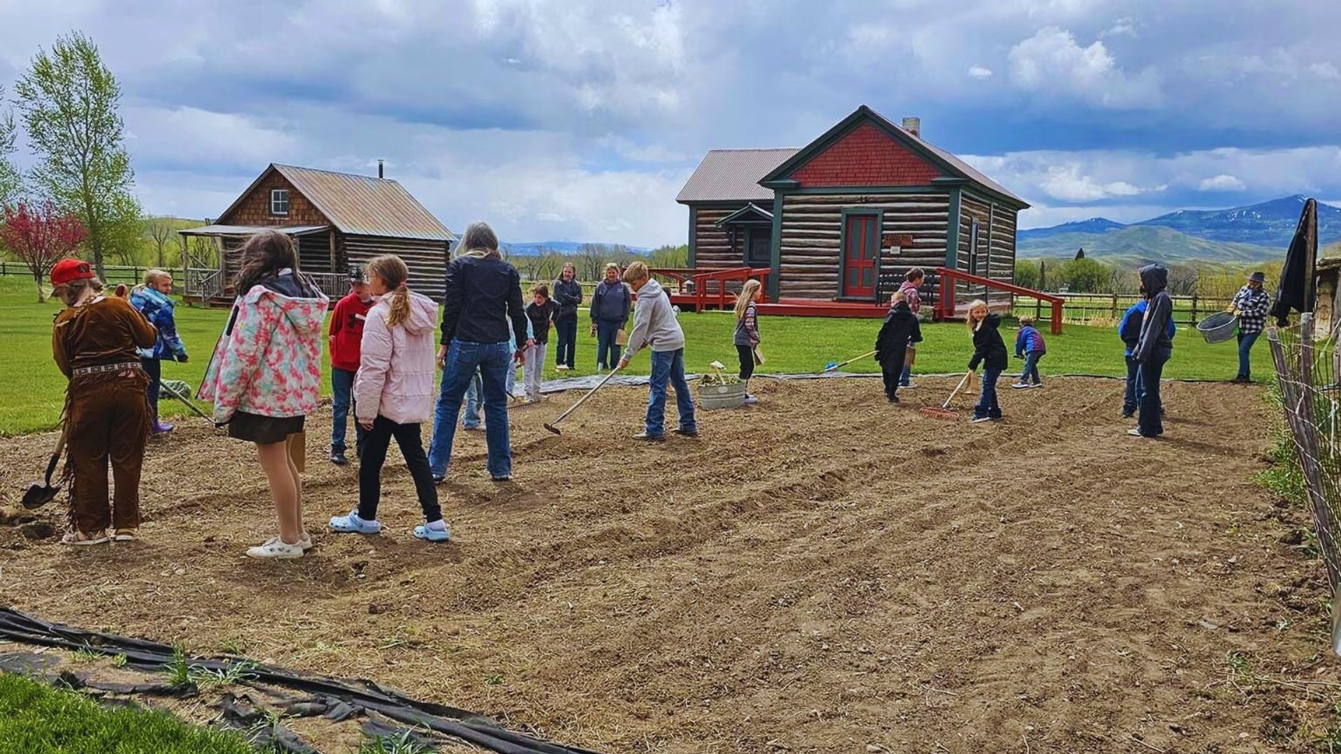 People are working in the soil in front of a rustic cabin, engaged in gardening or landscaping activities.