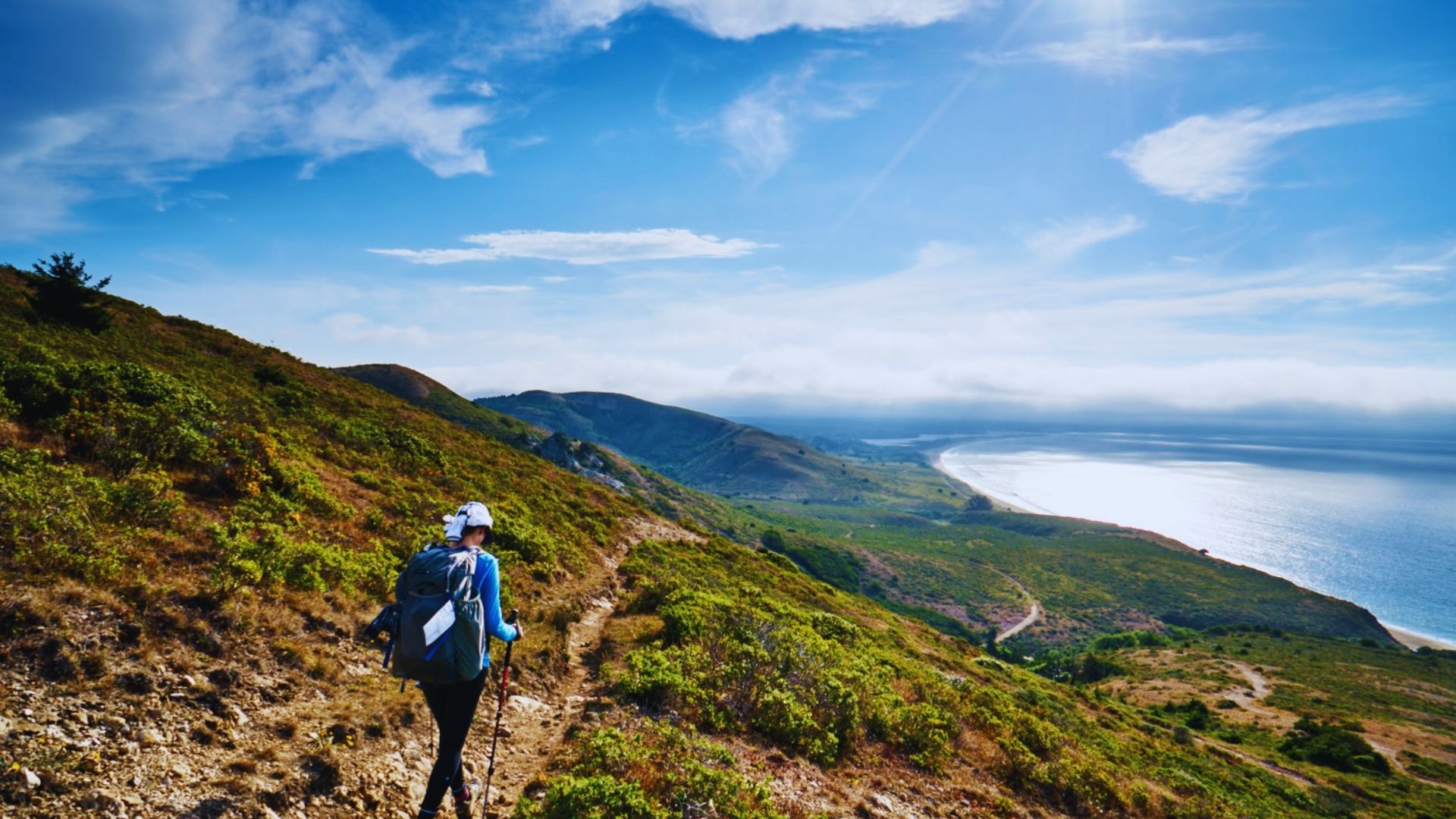 A man hiking uphill with a backpack, navigating rocky terrain on a mountain trail under a clear sky.