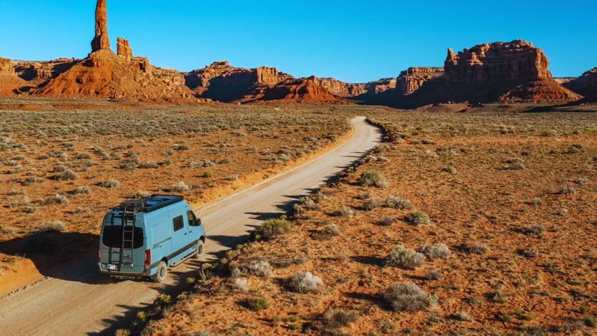 A van drives through the desert landscape of Monument Valley, Utah, surrounded by iconic red rock formations.