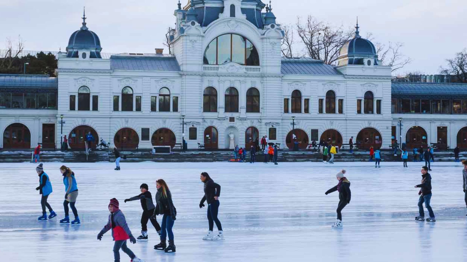 People skating on ice in front of a large, elegant building, enjoying a winter day outdoors.