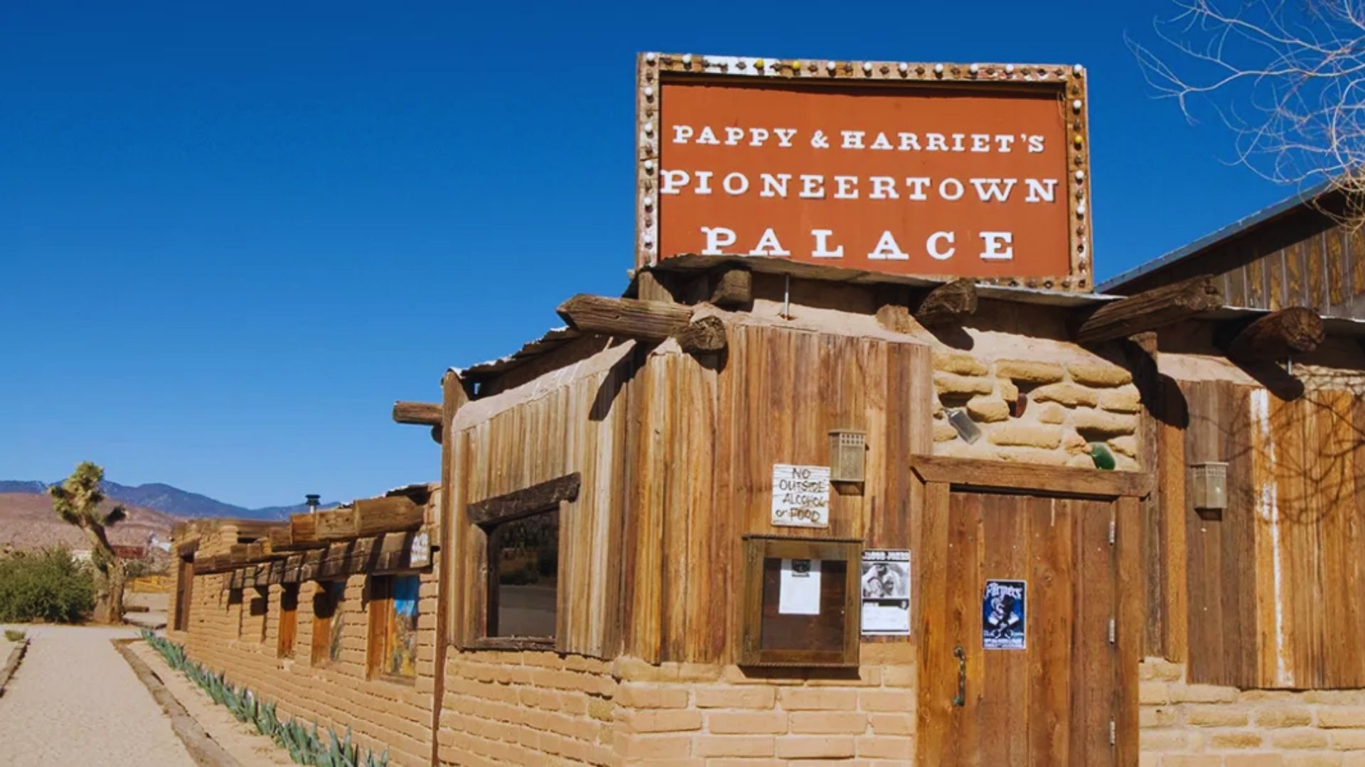 A sign for the Pioneer Town Palace stands prominently in a vast desert landscape under a clear blue sky.