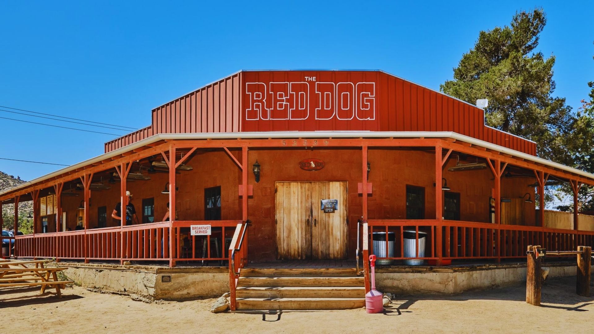 The Old Red Dog Saloon stands alone in a vast desert landscape under a clear blue sky.
