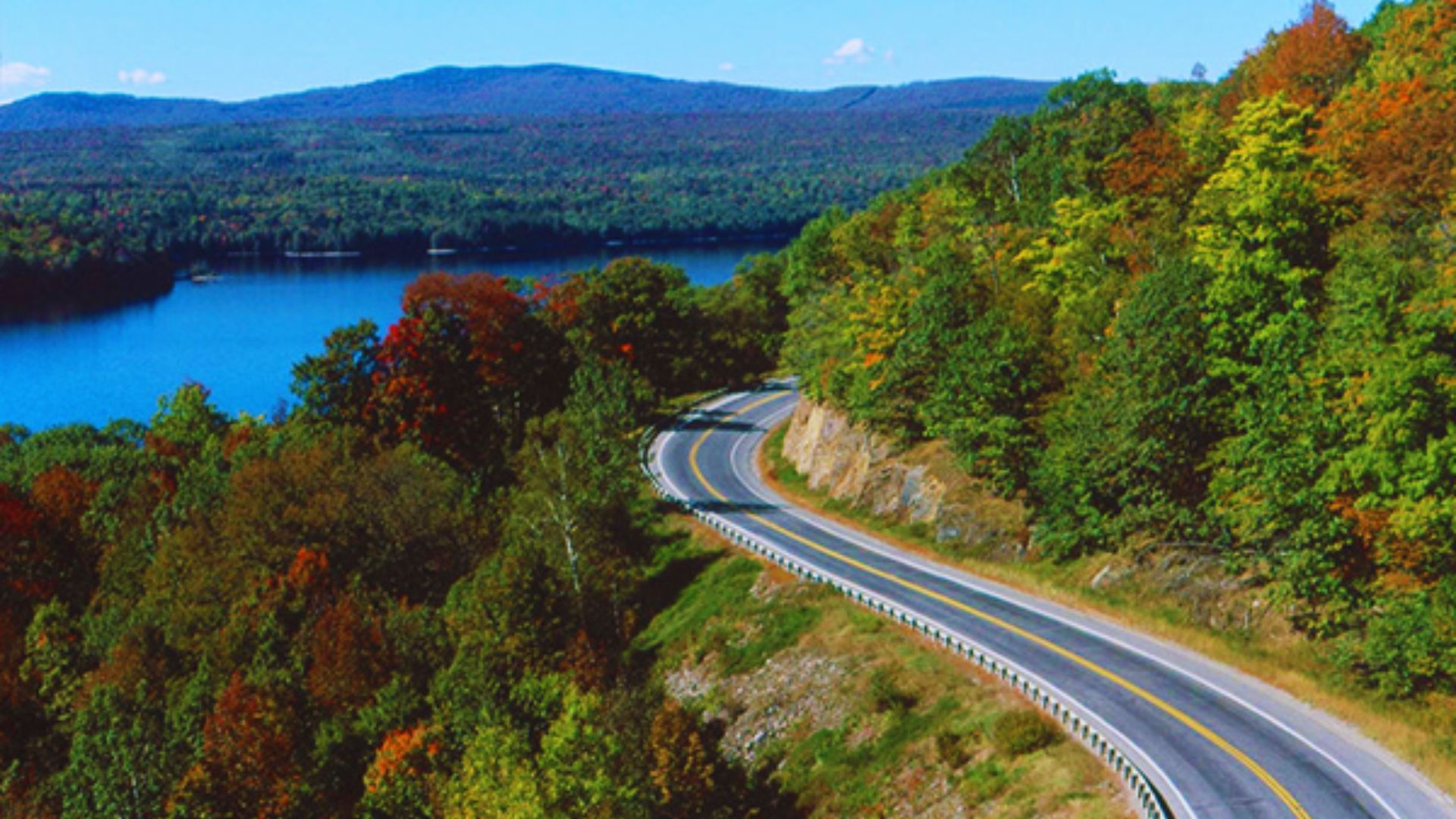 A winding road through mountains, surrounded by trees and a serene body of water, showcasing a picturesque landscape.