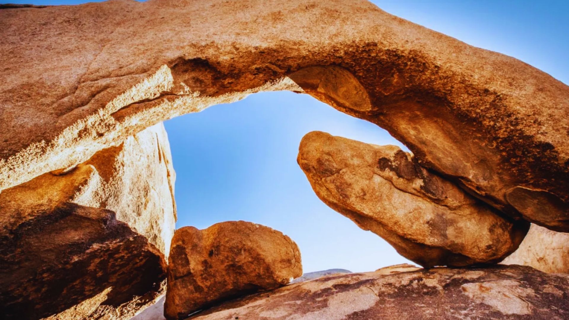 A natural rock arch stands in a desert landscape under a clear blue sky, showcasing the beauty of the arid environment.
