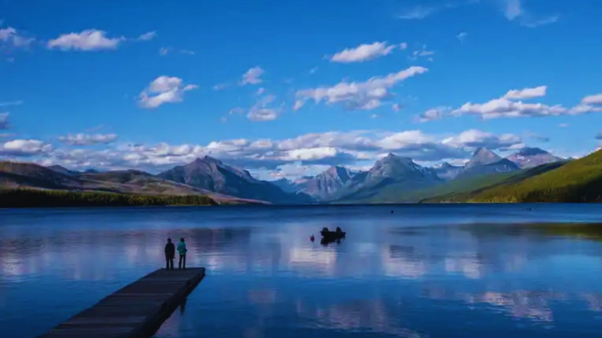 Two people stand on a dock, overlooking mountains in the background under a clear blue sky.
