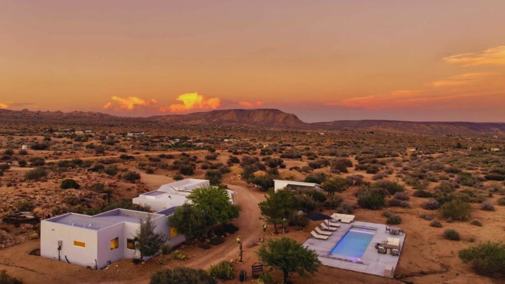 A sunset over a desert landscape featuring a white house and a swimming pool, casting warm hues across the scene.