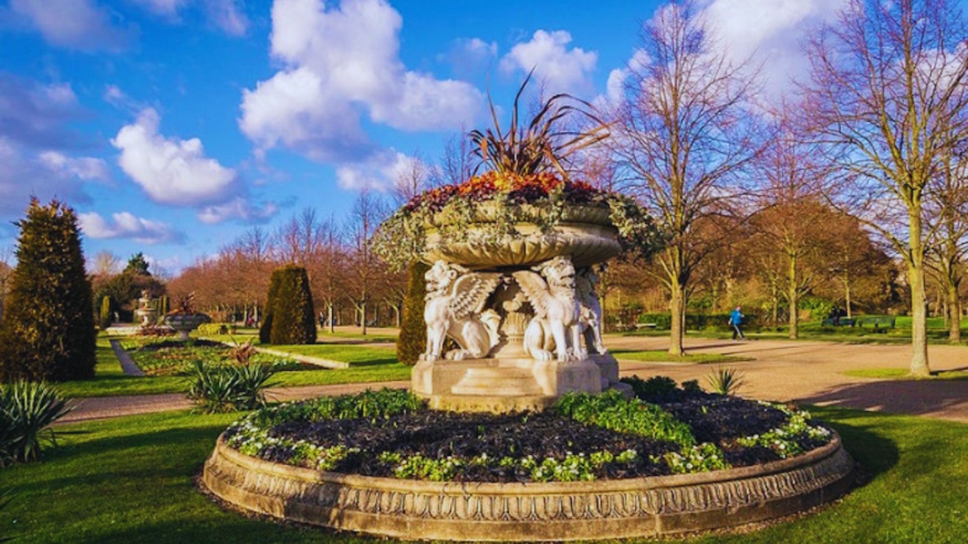A large fountain surrounded by trees and colorful flowers in a serene park setting.