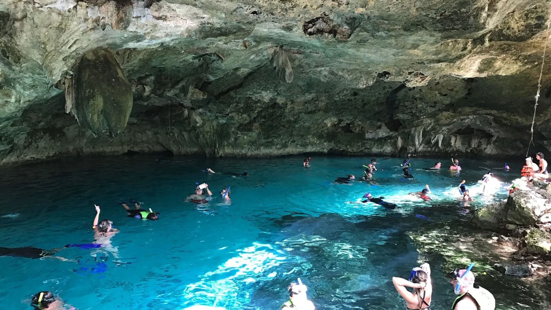 People swimming in the vibrant blue water of a cave, surrounded by rocky walls and natural light filtering in.