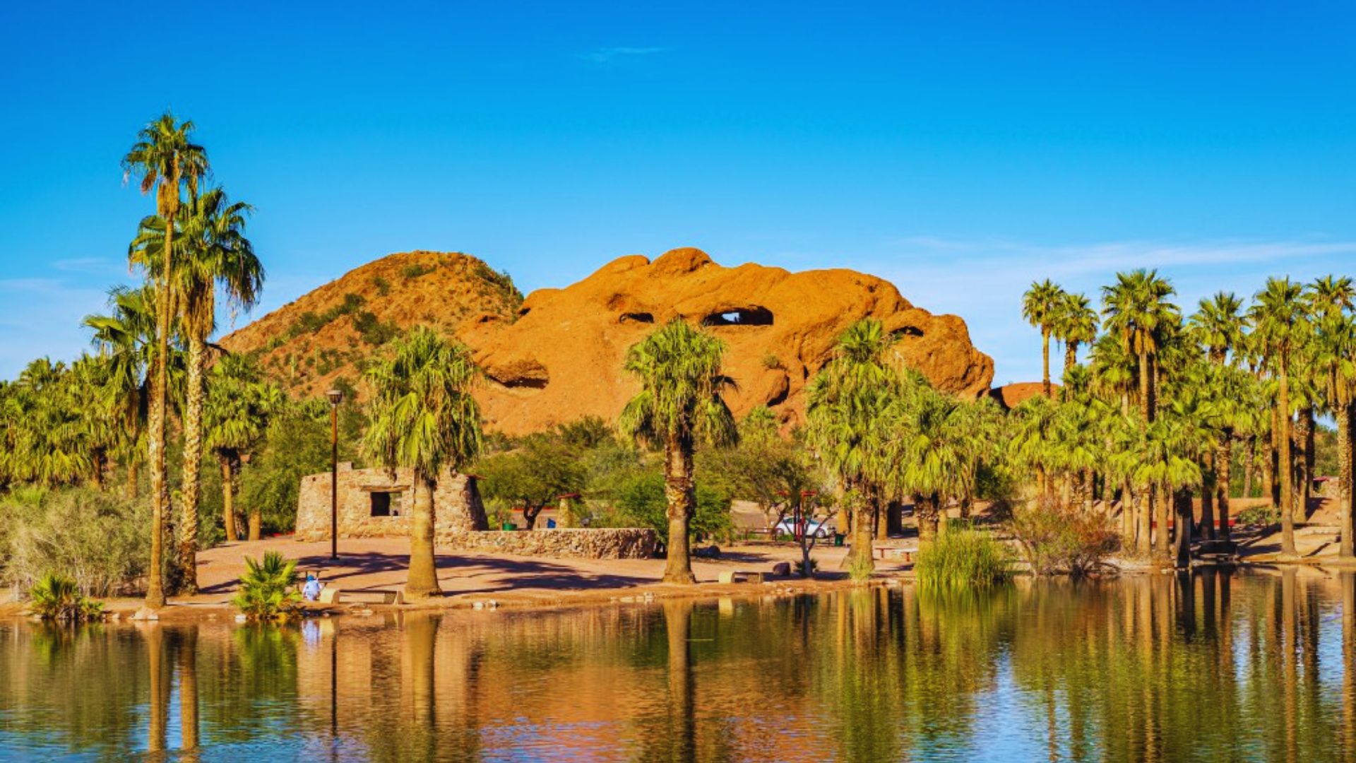 A serene lake bordered by lush palm trees and rugged rocks under a clear blue sky.