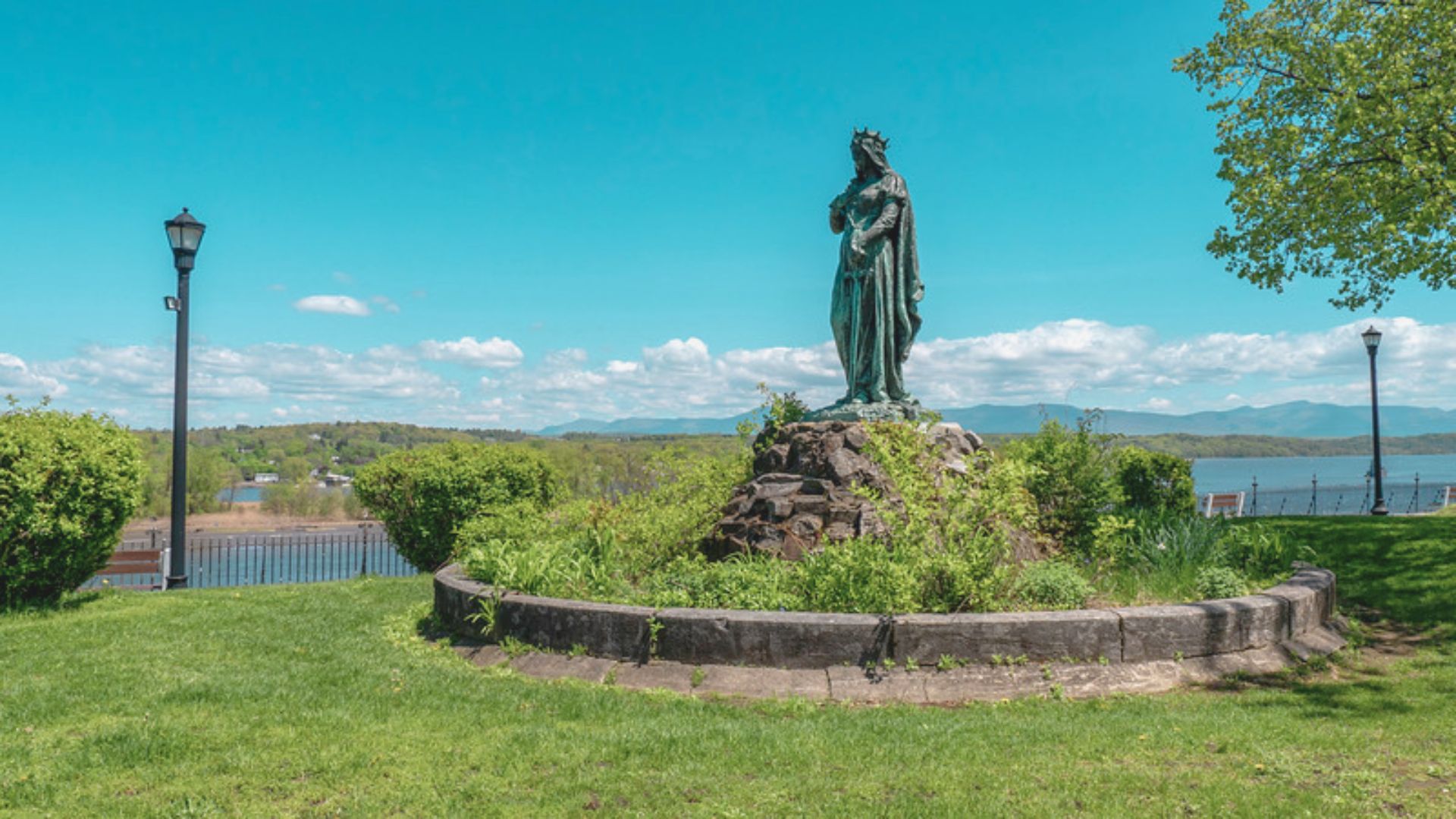 A statue of Mary stands gracefully in front of a serene lake, surrounded by lush greenery and a clear blue sky.
