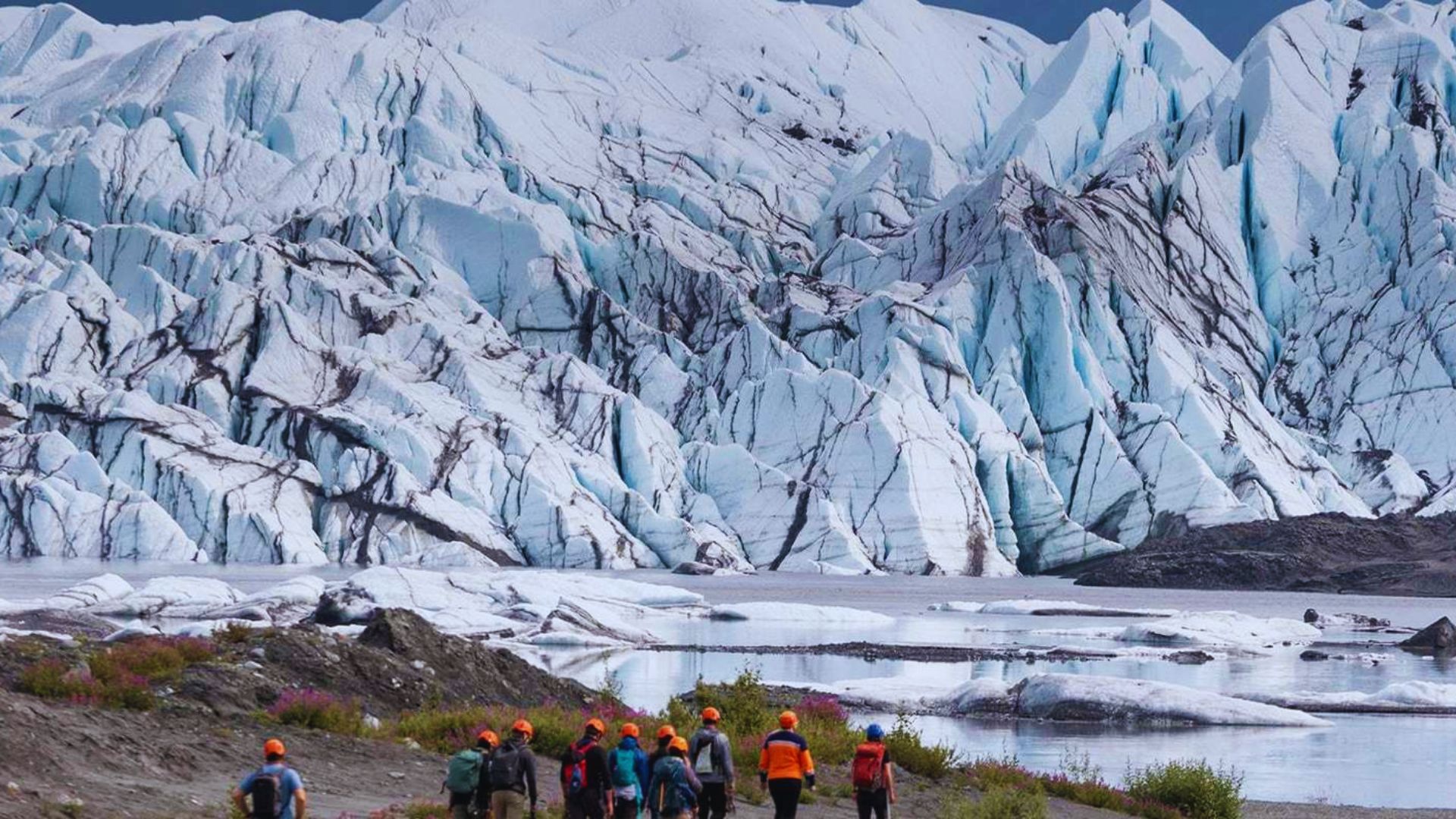 People walking along a scenic trail beside a glacier, surrounded by rugged mountains and clear blue skies.