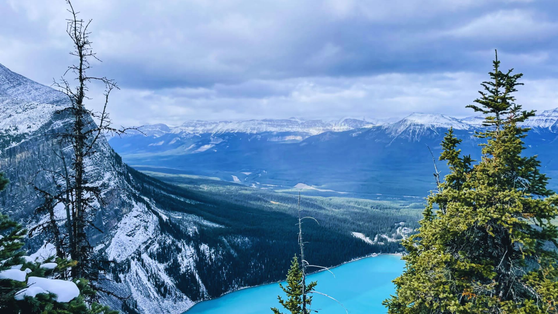 A panoramic view from a mountain peak, showcasing a serene lake surrounded by lush greenery and distant hills.