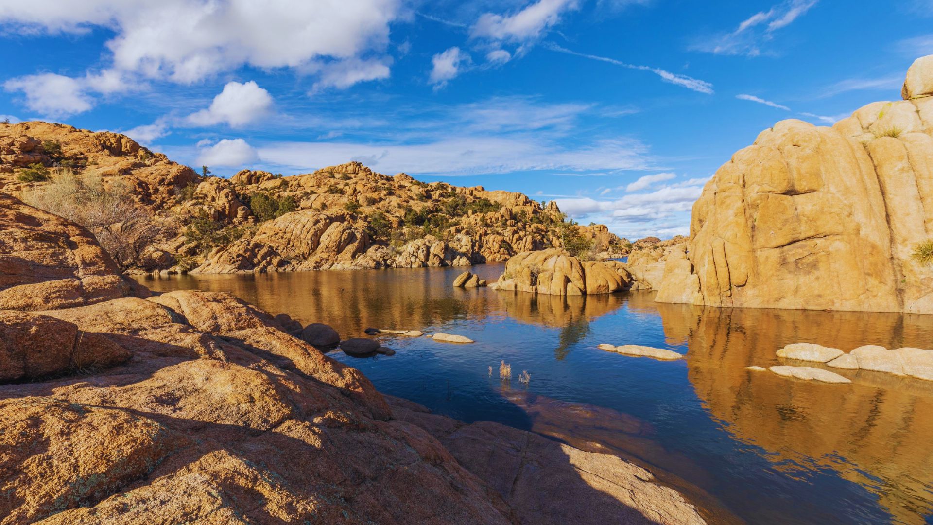 A rocky lake surrounded by desert terrain under a clear blue sky with fluffy white clouds.