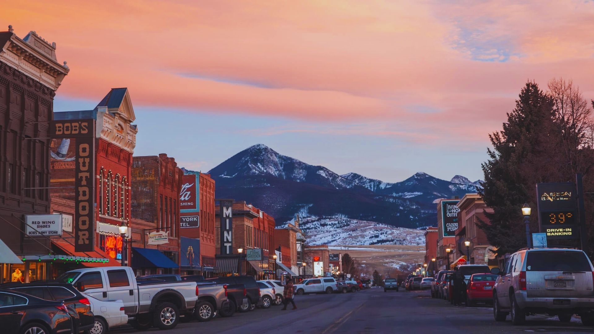 A bustling city street with tall buildings, framed by majestic mountains in the background under a clear blue sky.
