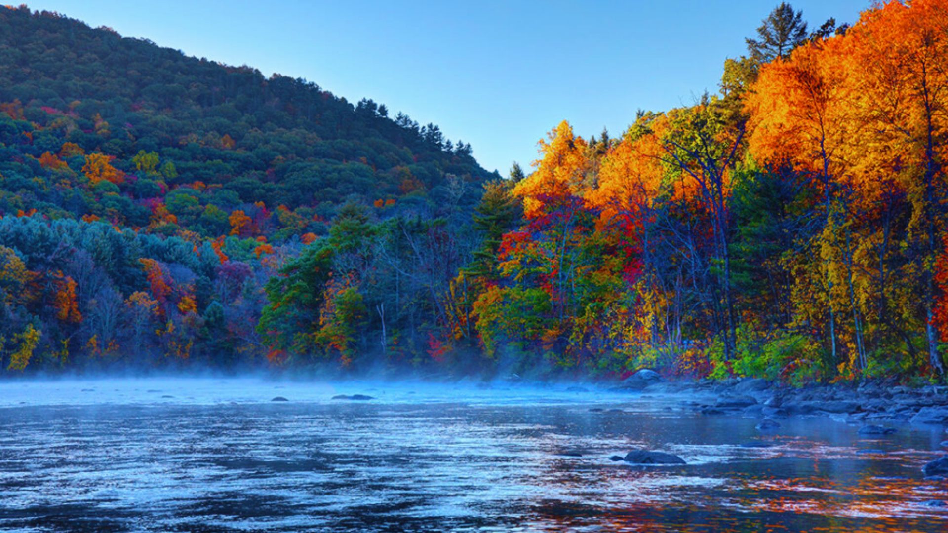 Autumn mist envelops a serene river flowing through majestic mountains, creating a tranquil and picturesque landscape.