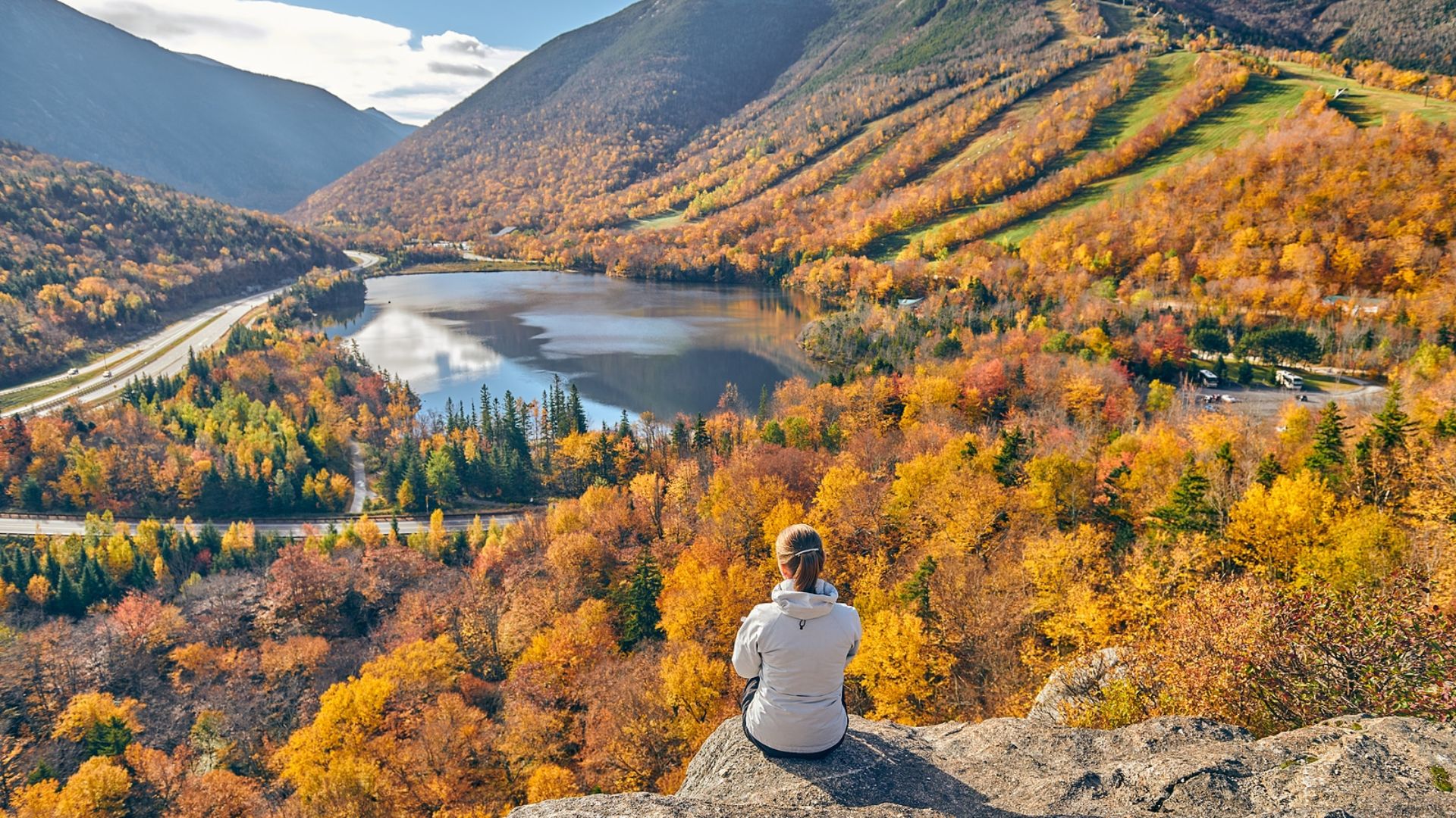 A woman sits on a rock, gazing at a serene lake surrounded by majestic mountains in the background.