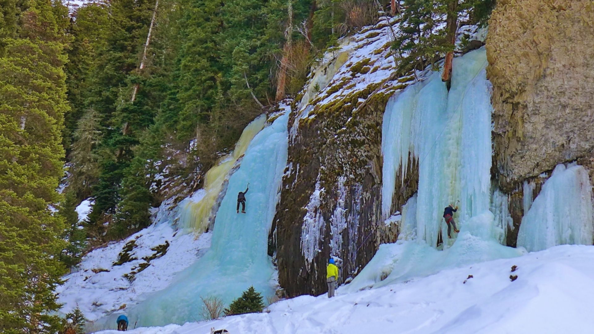 A climber ascends a steep ice wall in a rugged mountain landscape, surrounded by snow and rocky peaks.