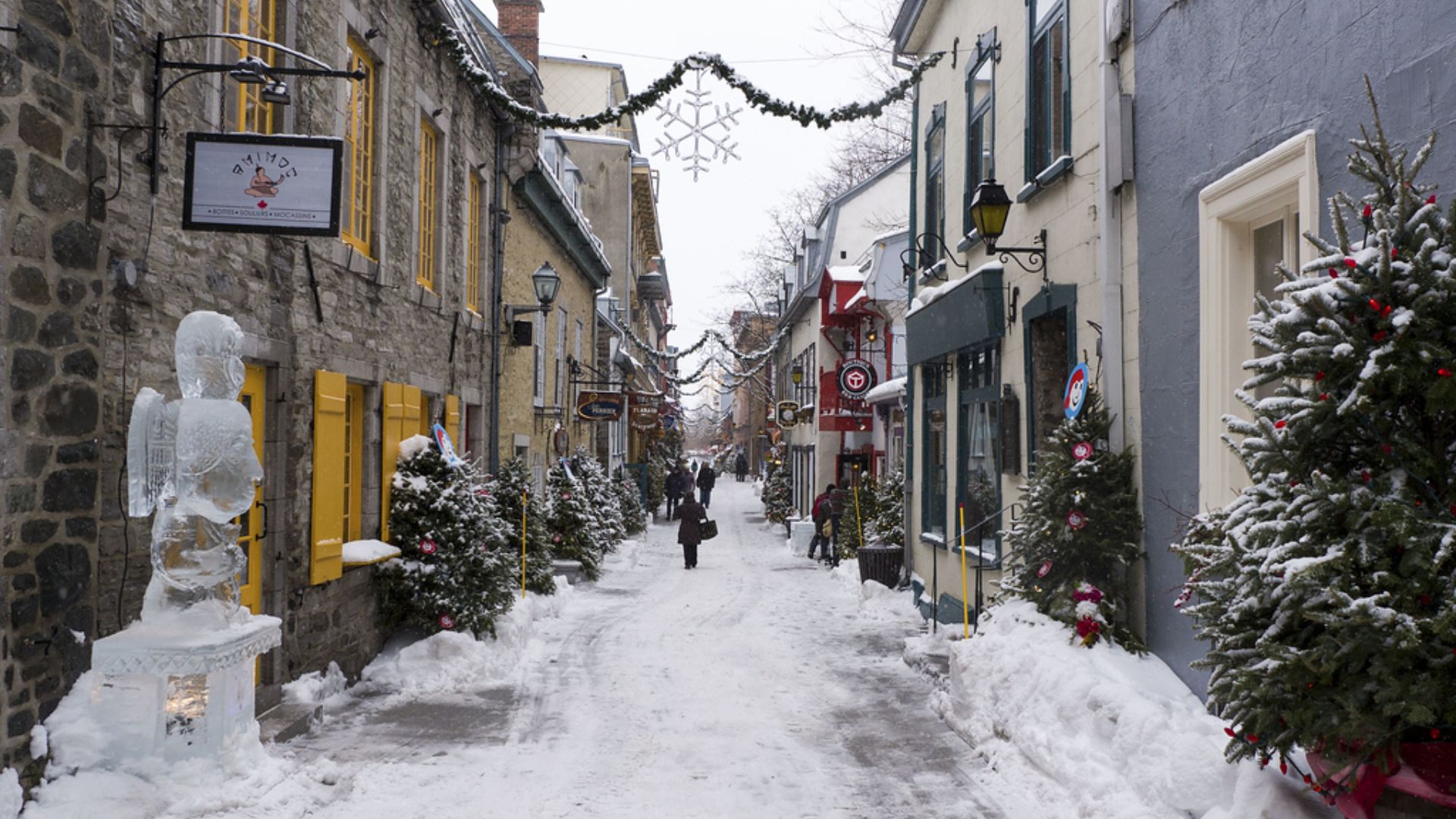 A snowy street scene featuring a decorated Christmas tree and a statue, creating a festive winter atmosphere.