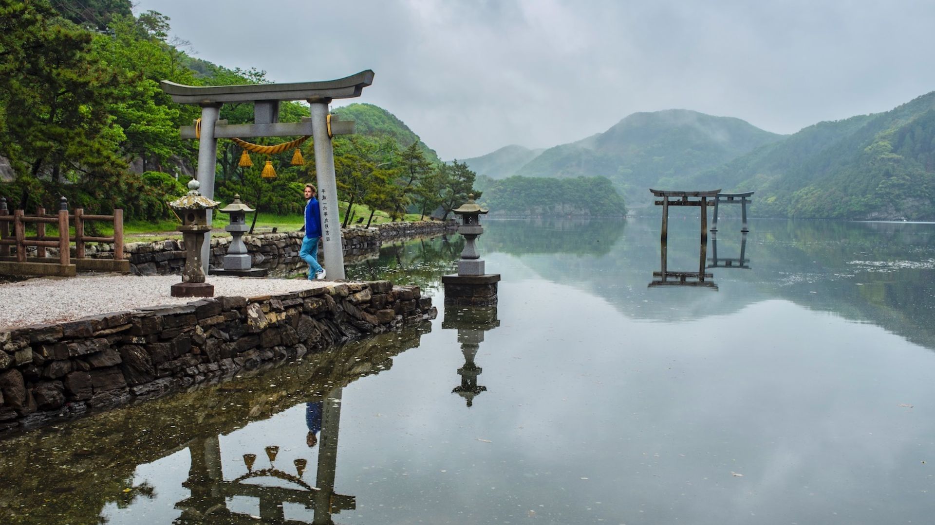 A red torii gate stands prominently in the center of a serene lake, surrounded by calm waters and distant trees.