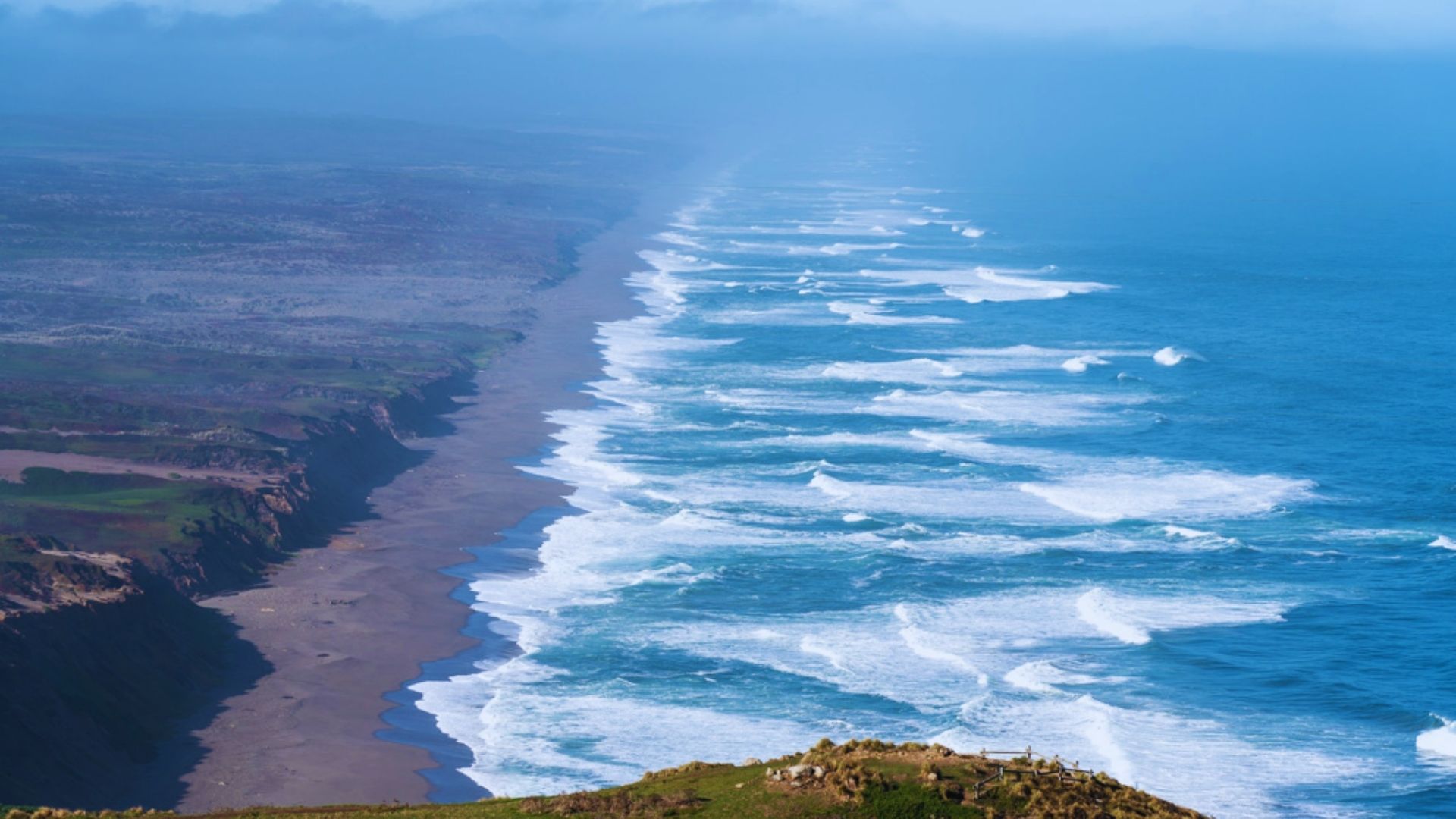 A scenic view of the ocean and coastline from a hill, showcasing waves crashing against the shore under a clear sky.