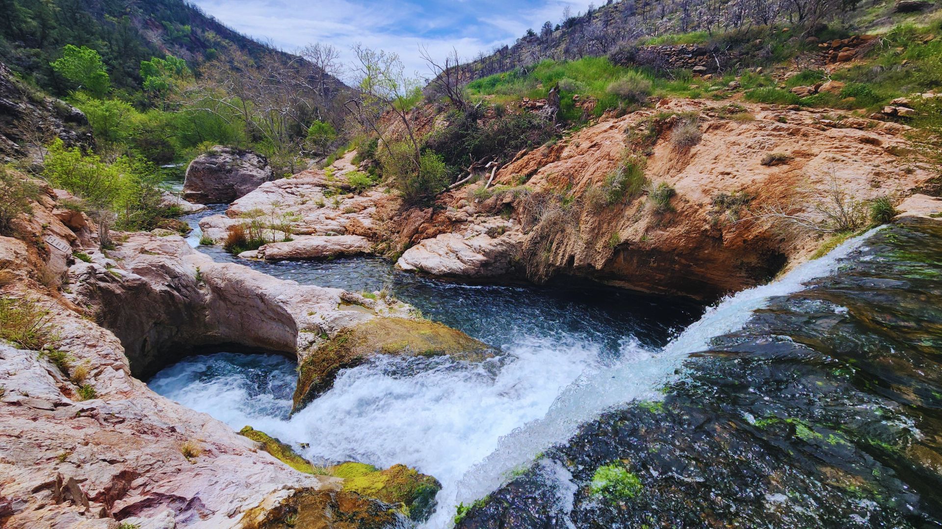 A river flows through a rocky canyon, with clear water cascading over stones and surrounded by steep canyon walls.