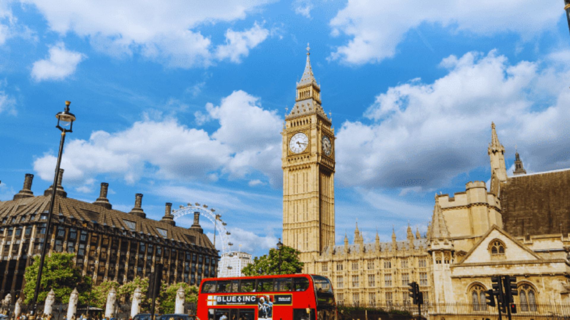 A double-decker bus travels down a street with a clock tower visible in the background.