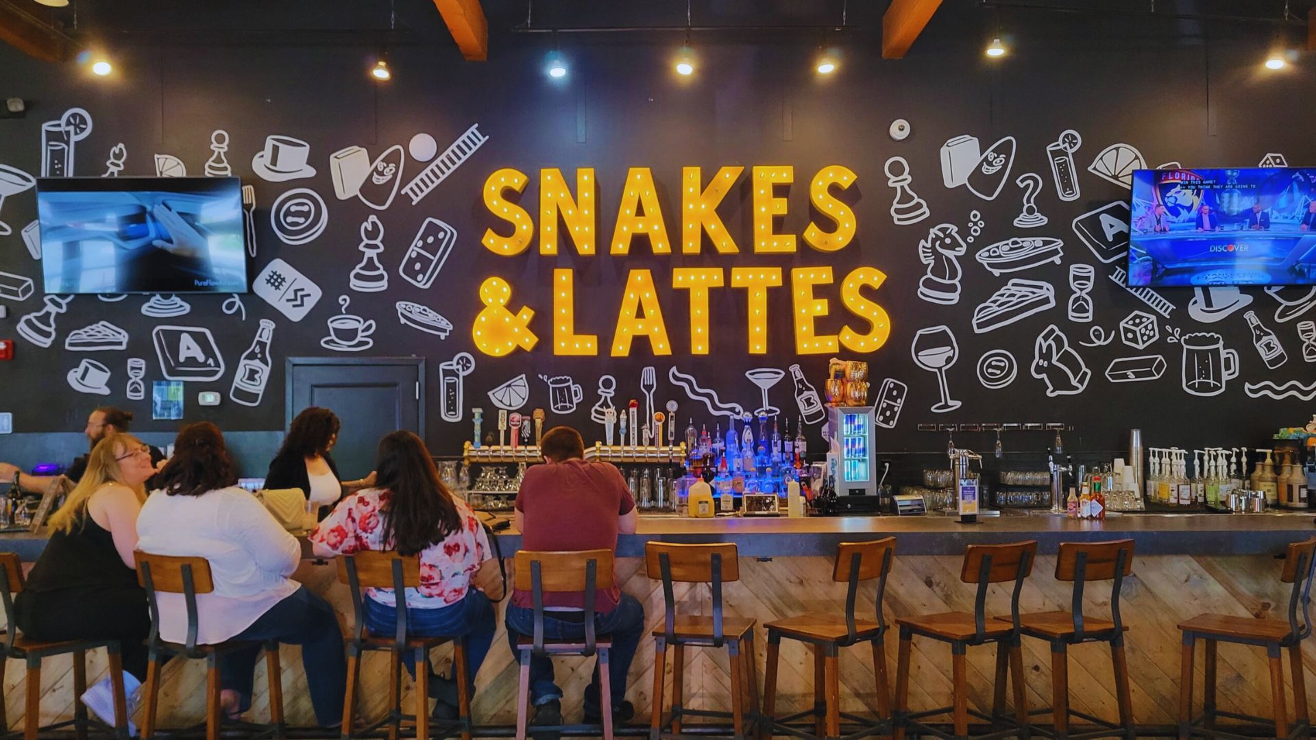 A cozy bar featuring snakes and lattes, with patrons enjoying drinks and games in a vibrant, welcoming atmosphere