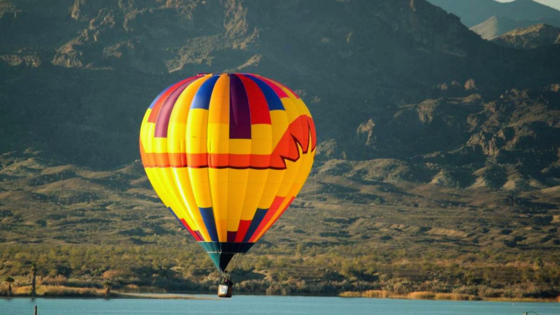 A vibrant hot air balloon soars above a serene lake, reflecting colorful patterns on the water's surface.