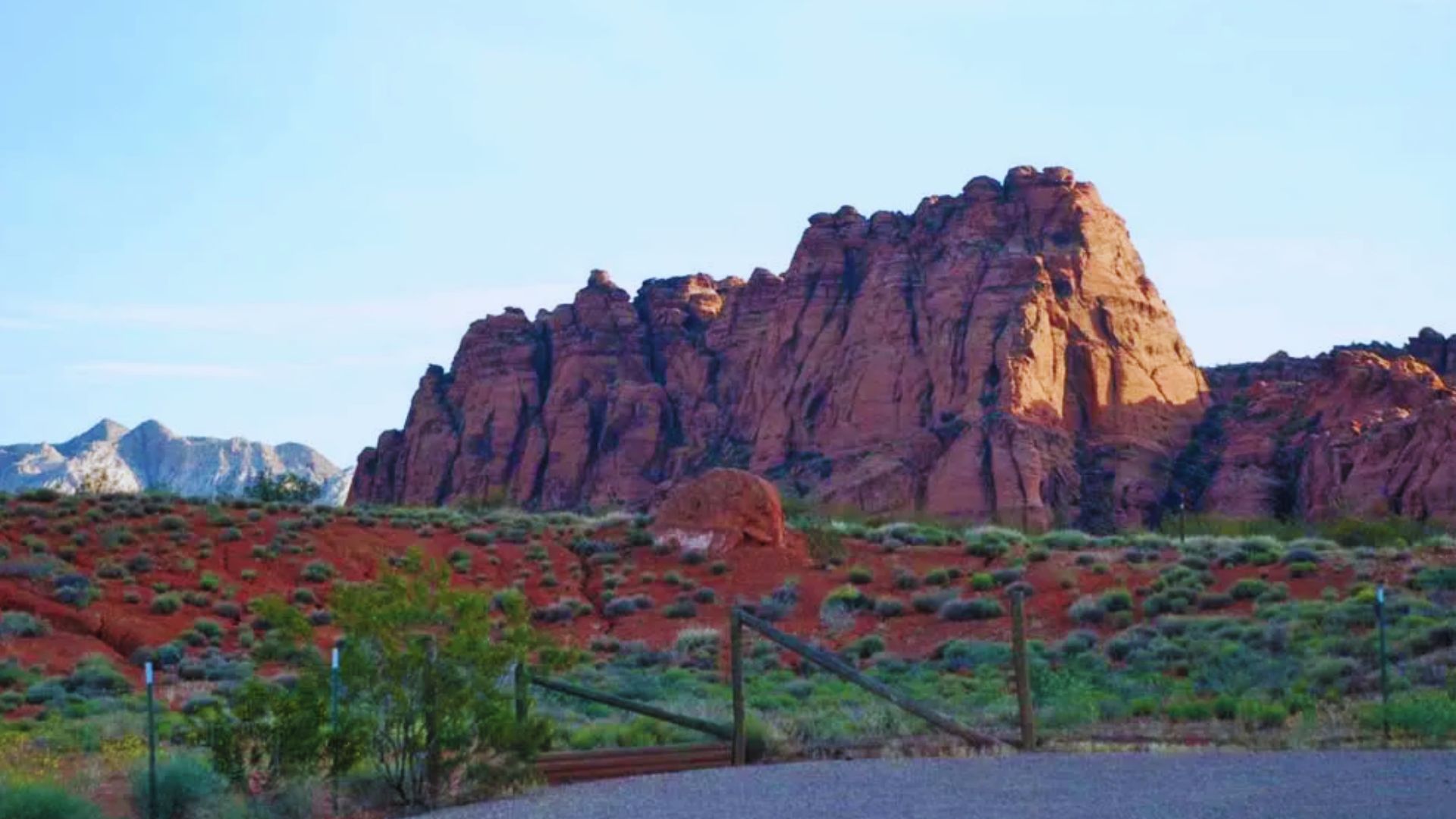 A striking red rock formation in the desert, bordered by a wooden fence under a clear blue sky.