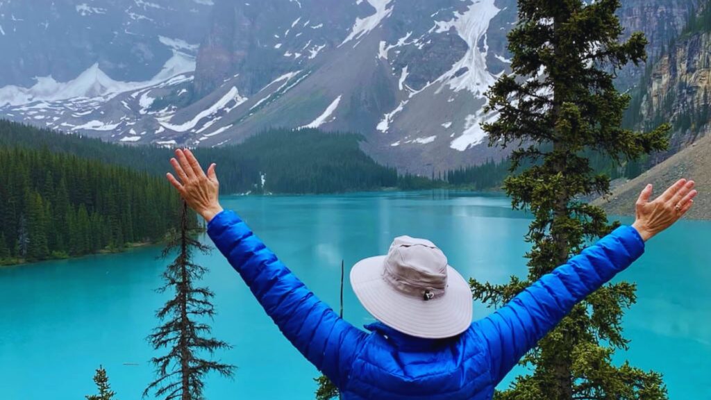 A person stands with arms outstretched in front of a serene mountain lake, surrounded by lush greenery and peaks.