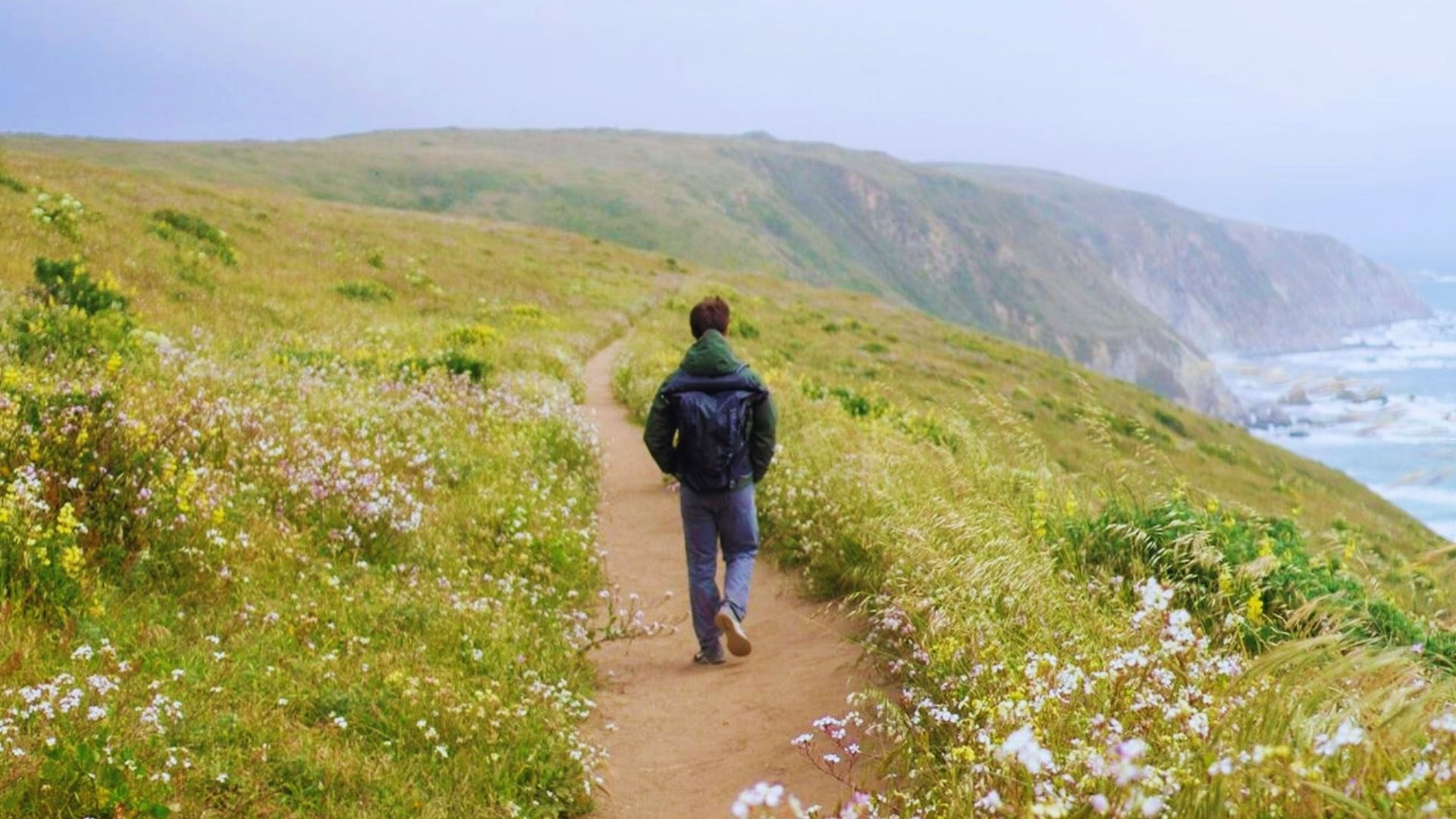 A man walks along a seaside path, with waves gently crashing nearby and a bright sky overhead.