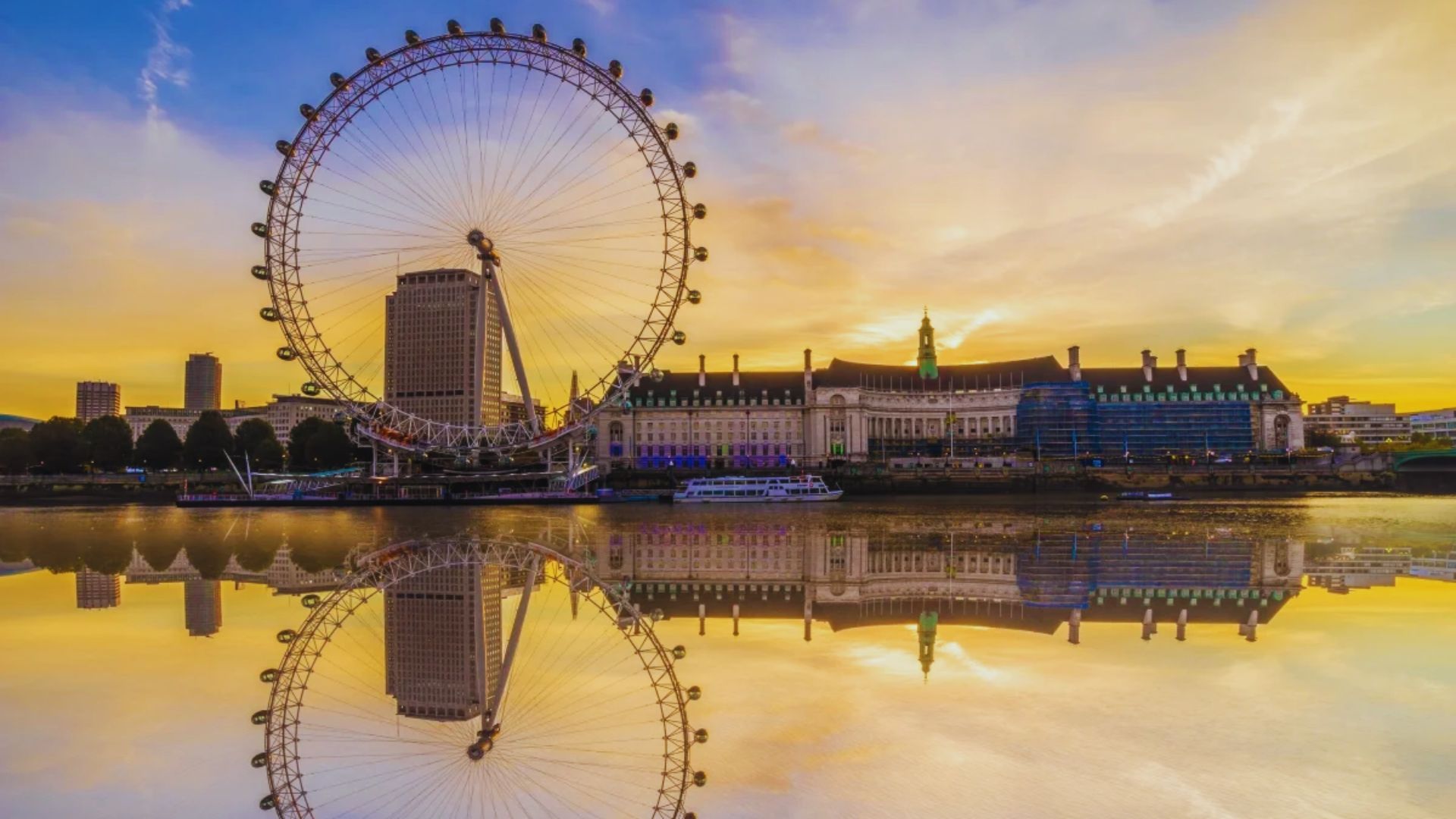 The London Eye silhouetted against a vibrant sunset sky, casting reflections on the River Thames below.