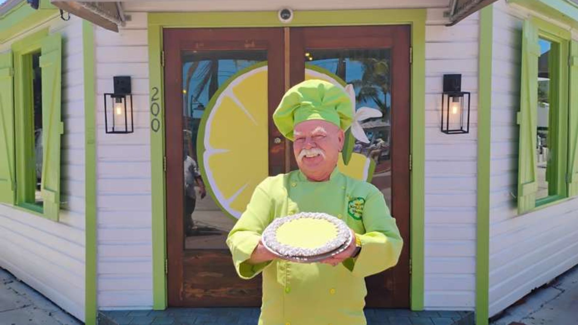 A chef in a white uniform presents a pie, showcasing his culinary creation with a cheerful expression.