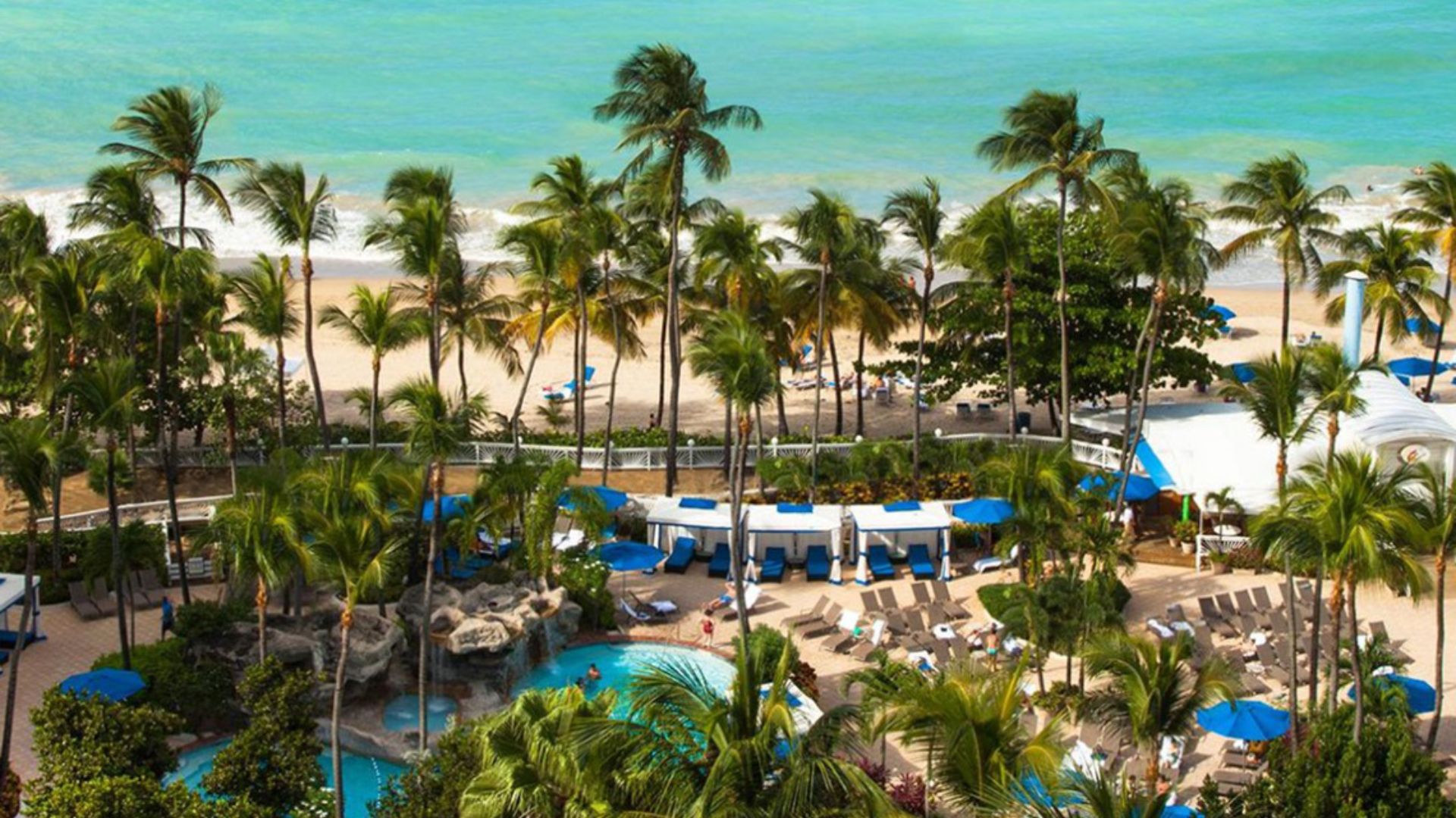 Aerial view of a beach and pool area, showcasing sandy shores and a sparkling blue pool surrounded by lounge chairs.