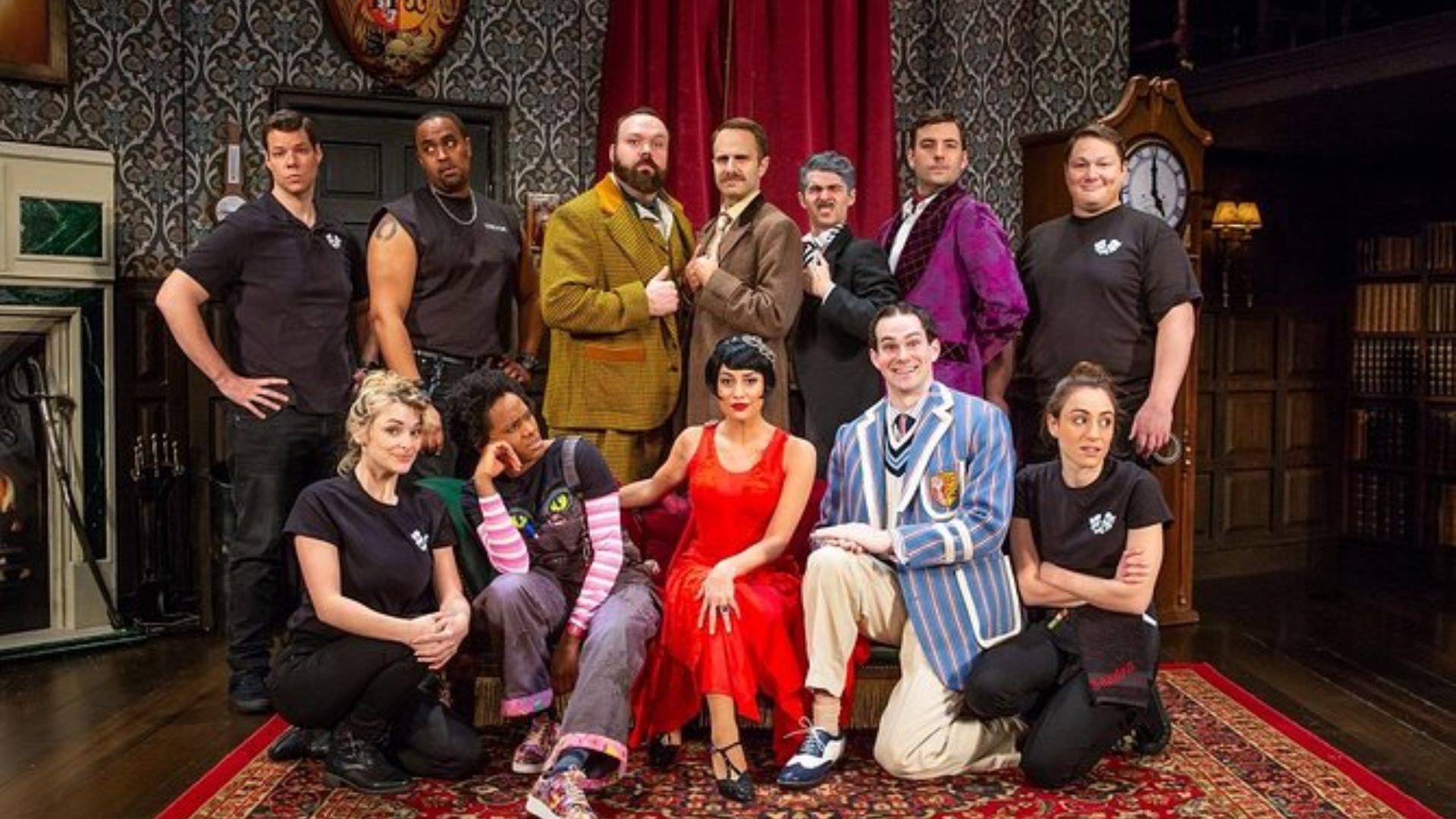 The musical cast stands together, featuring a woman in the middle, showcasing their colorful costumes and joyful expressions.