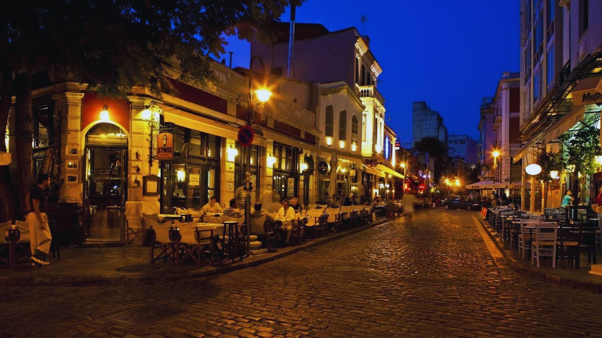 A cobblestone street at night, featuring outdoor tables and chairs illuminated by soft lighting.