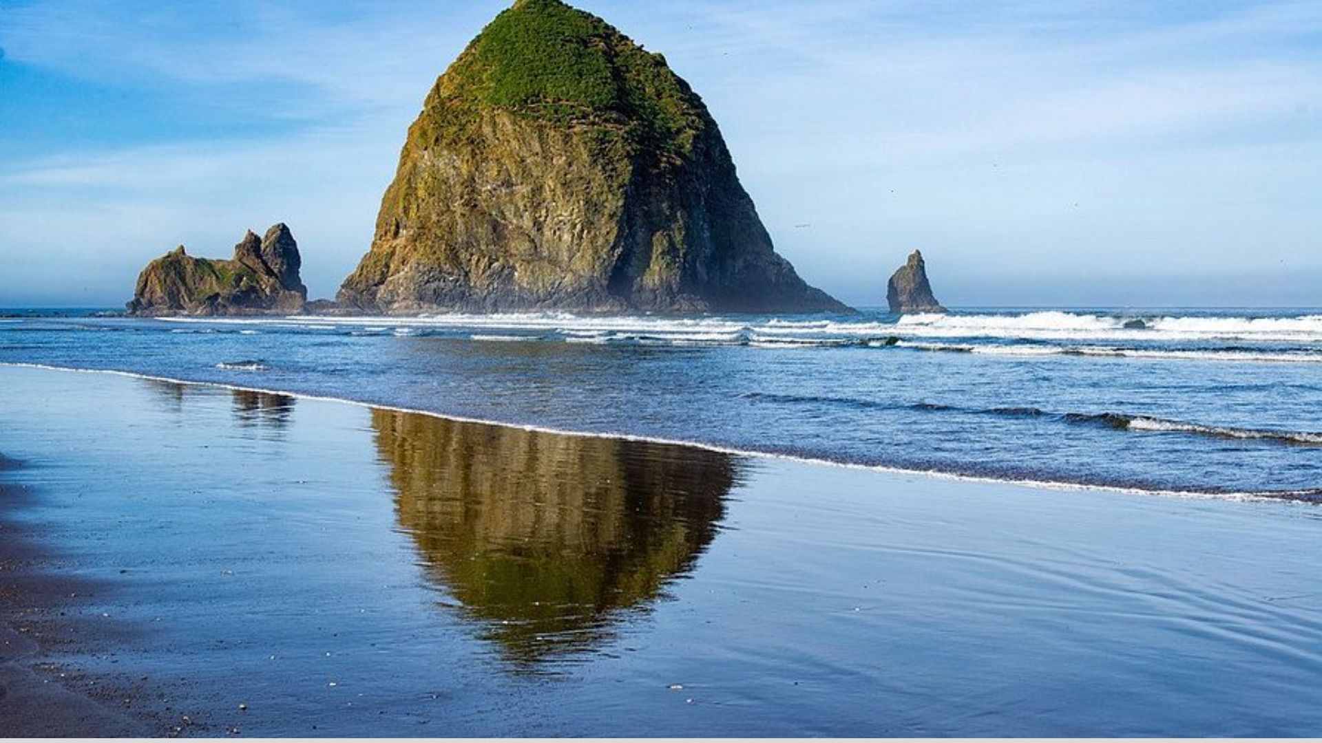 . Haystack Rock rises majestically at Cannon Beach, Oregon, surrounded by sandy shores and gentle ocean waves.