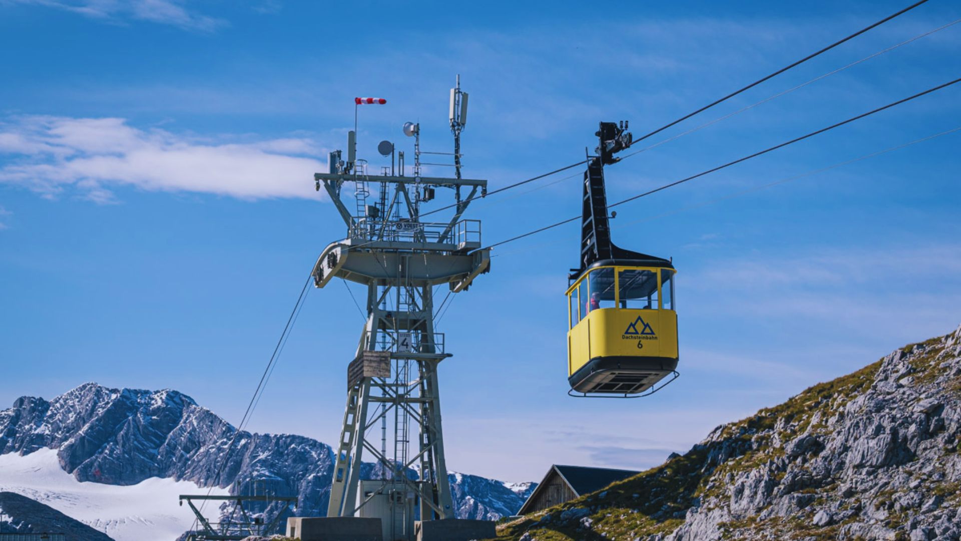 A cable car ascends a mountain, surrounded by lush greenery and rocky terrain under a clear blue sky.
