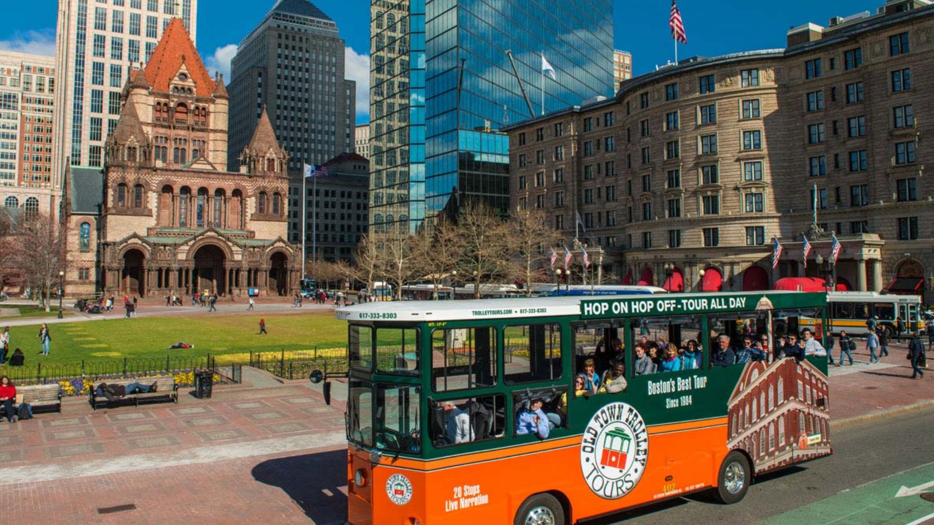 A trolley bus travels down a city street, flanked by tall buildings in the background under a clear blue sky.