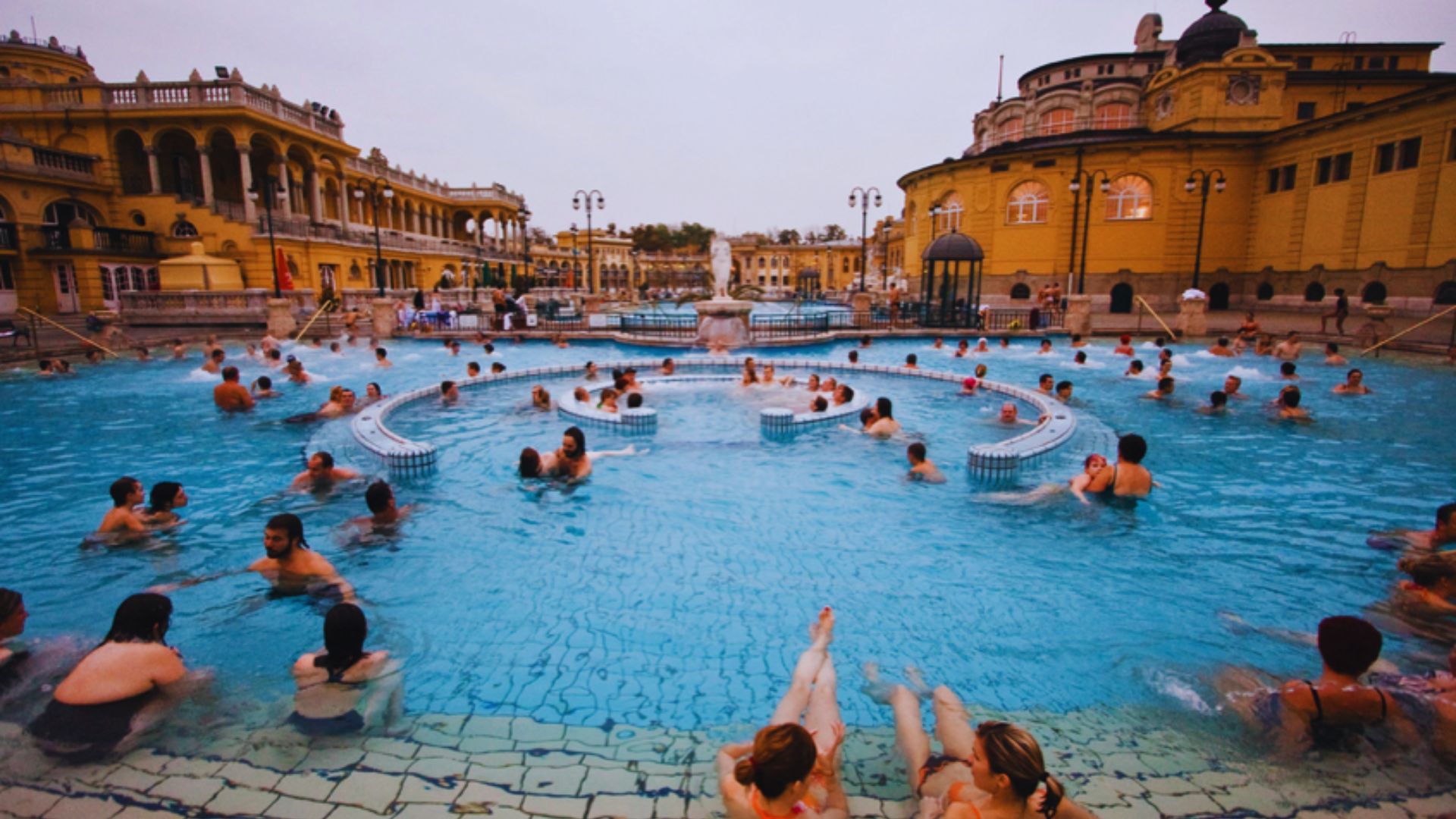 People relaxing and enjoying the thermal hot springs in Budapest, surrounded by historic architecture and lush greenery.