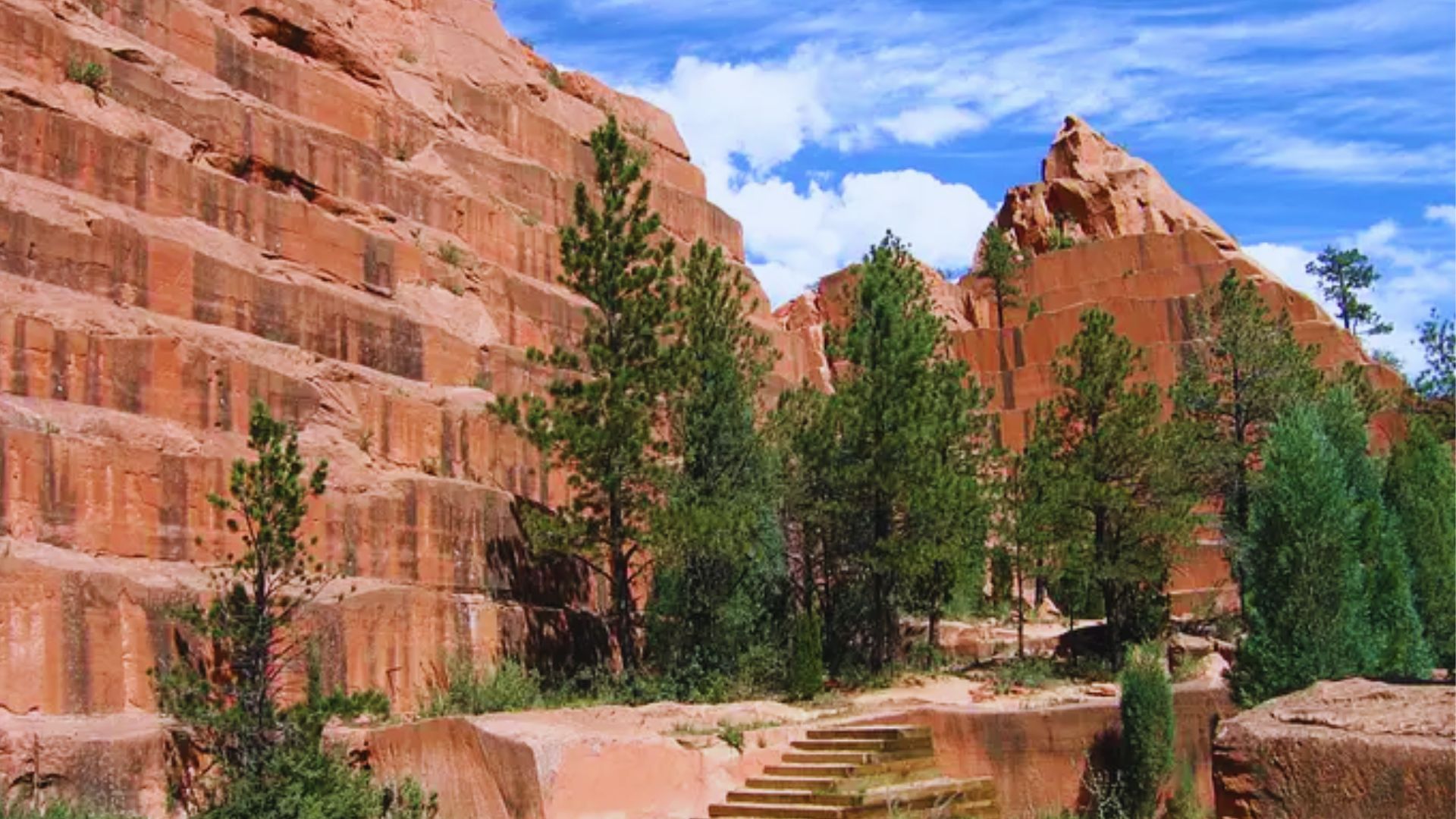 A large rock formation with stairs ascending to its summit, surrounded by greenery and a clear blue sky.