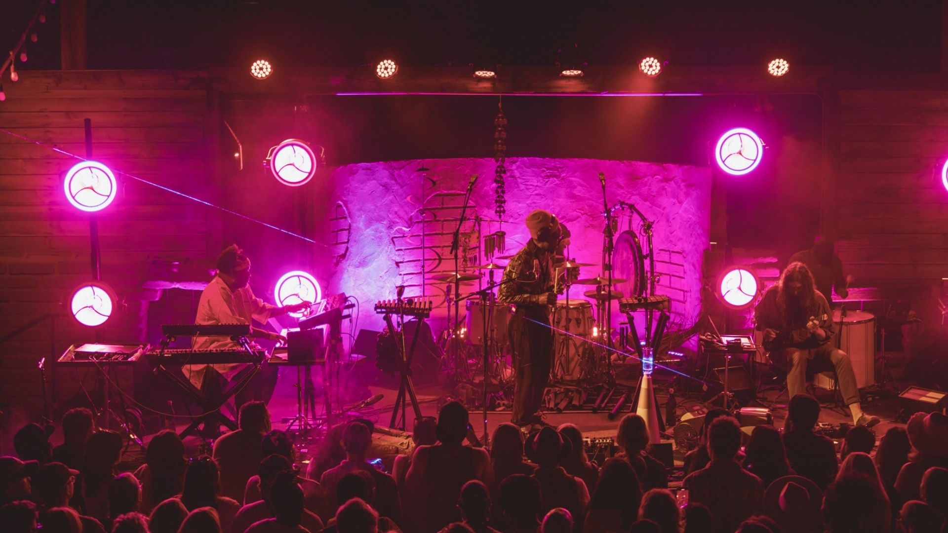 A band performs on stage, illuminated by vibrant pink lights, creating an energetic atmosphere.