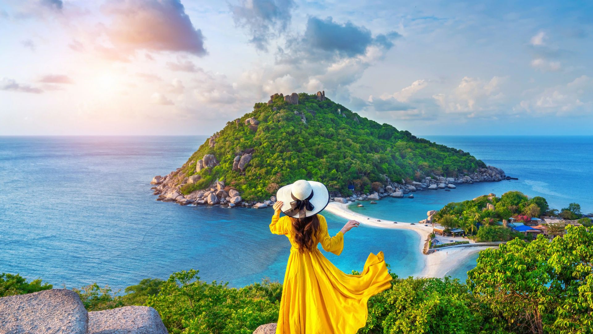 A woman wearing a yellow dress and hat overlooks the ocean from a hilltop.