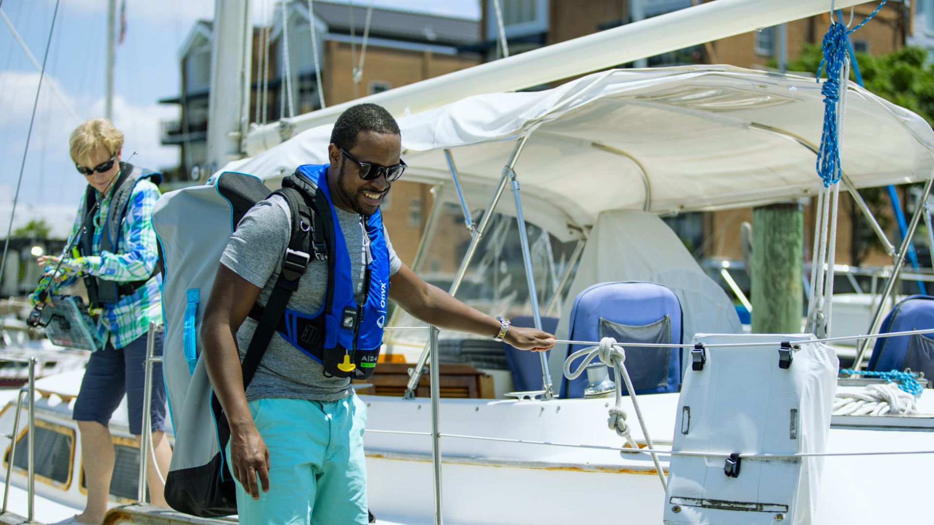 A man in a blue shirt and shorts stands on the deck of a sailboat, enjoying the view of the water.