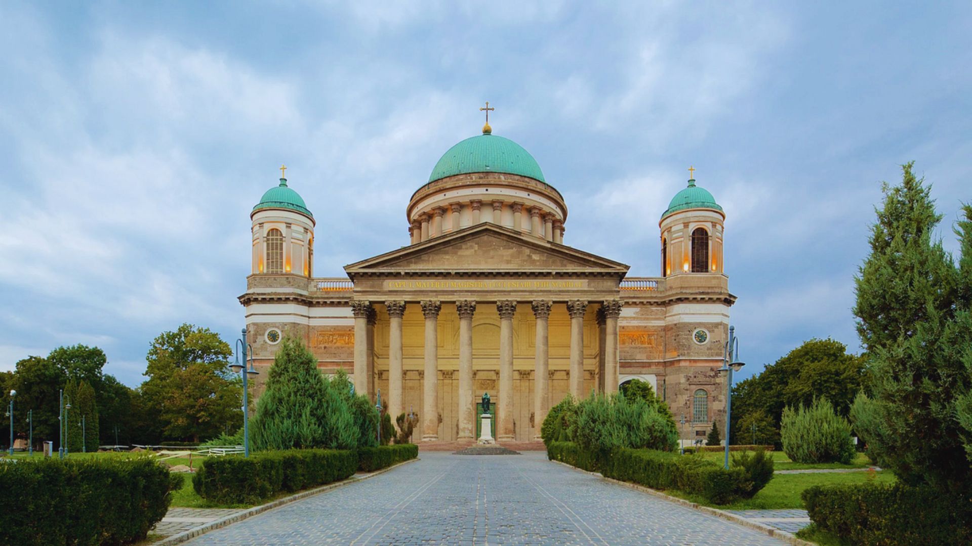 Front view of a neoclassical cathedral with a central dome and two green-domed towers, surrounded by greenery under a cloudy blue sky.