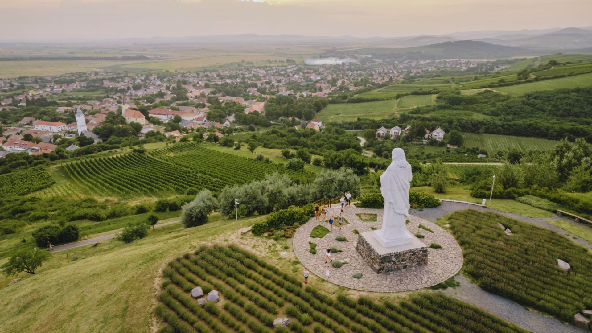 Aerial view of a large white statue on a pedestal surrounded by gardens and pathways. The statue overlooks a village with fields and hills in the distance, under a soft sunset sky.
