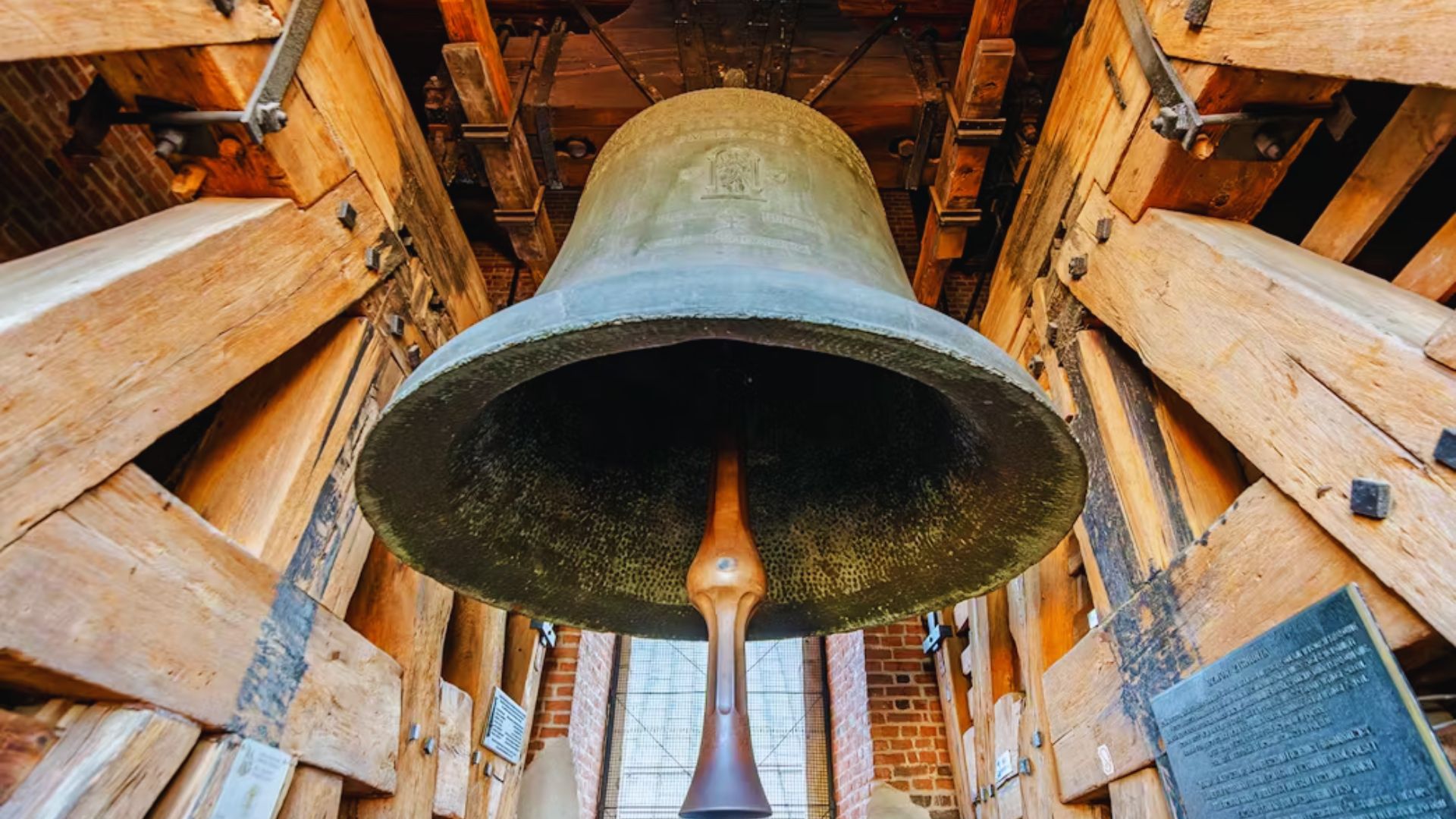 A large bell hangs inside a building with rustic wooden walls, showcasing its size and craftsmanship.