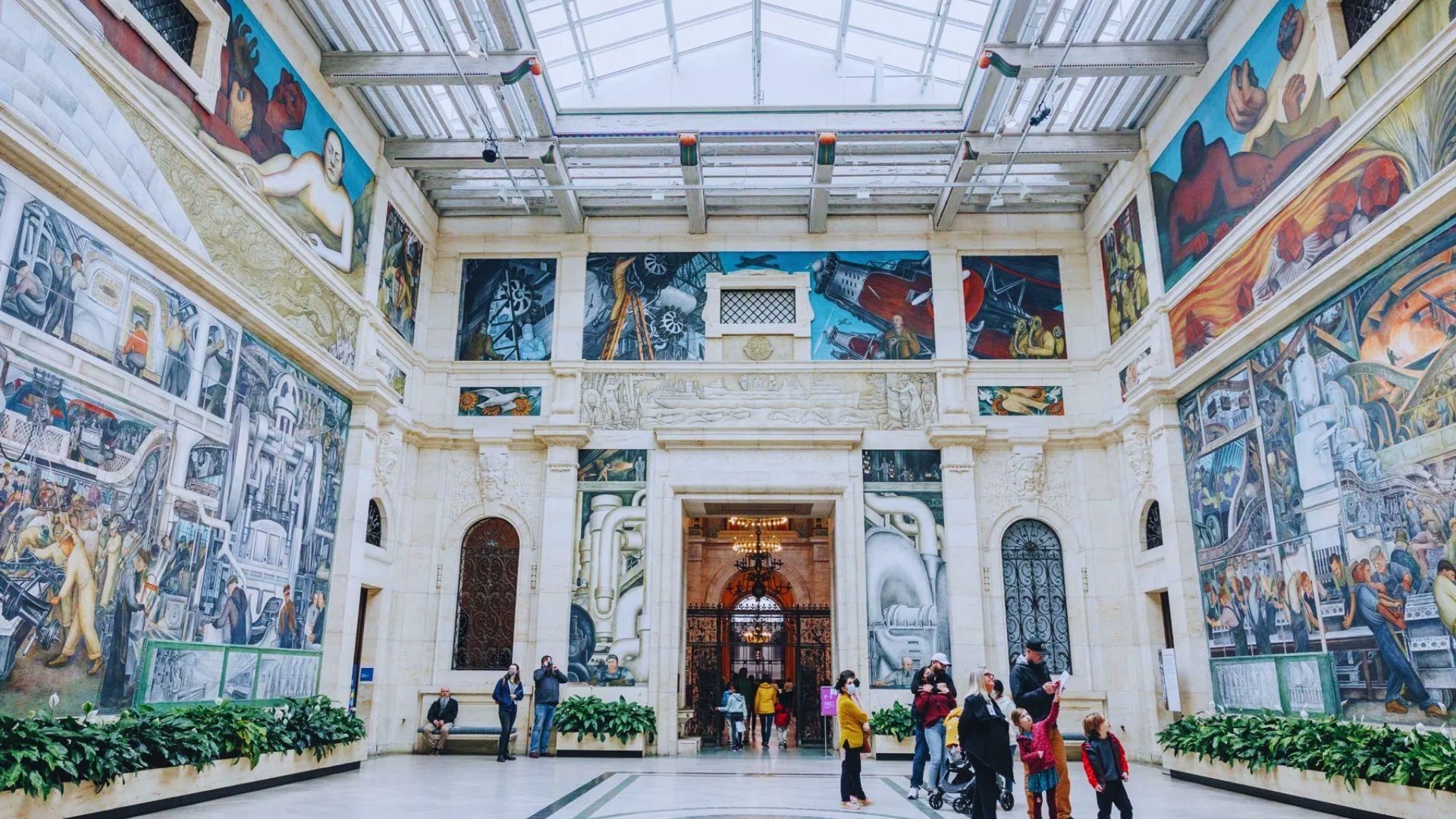 People walk through a spacious building adorned with various paintings on the walls.